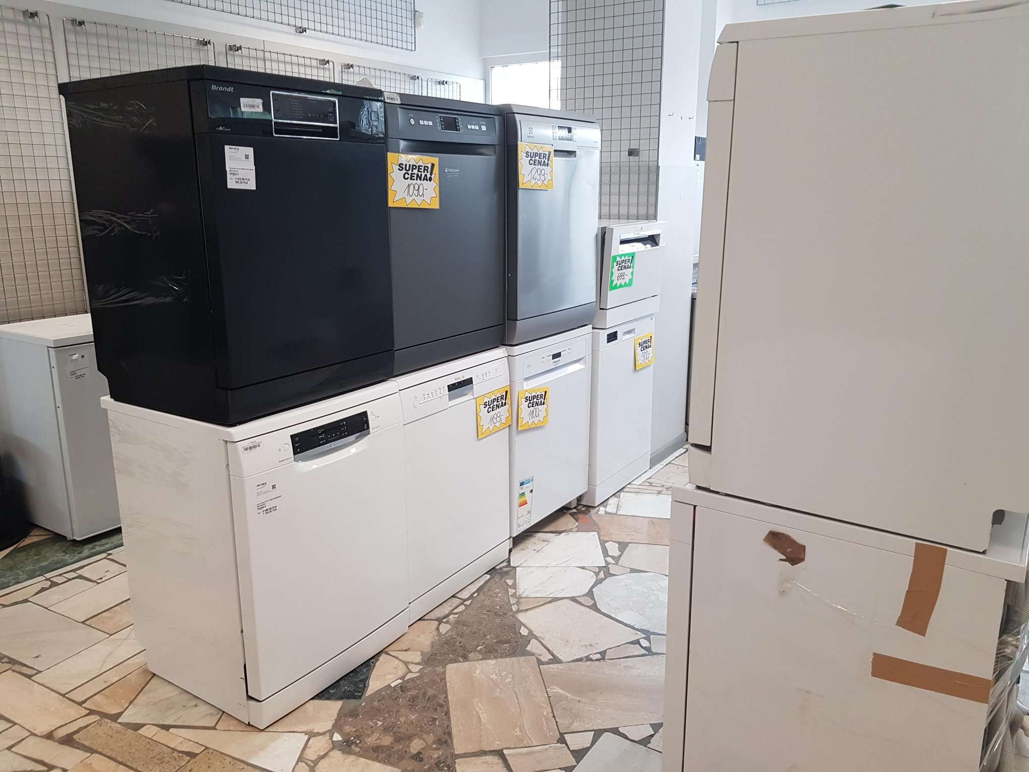 Rząd nowych zmywarek w sklepie, różne modele i kolory, z etykietami cenowymi 'SUPER CENA' na każdym urządzeniu, ustawione na kafelkowej podłodze.