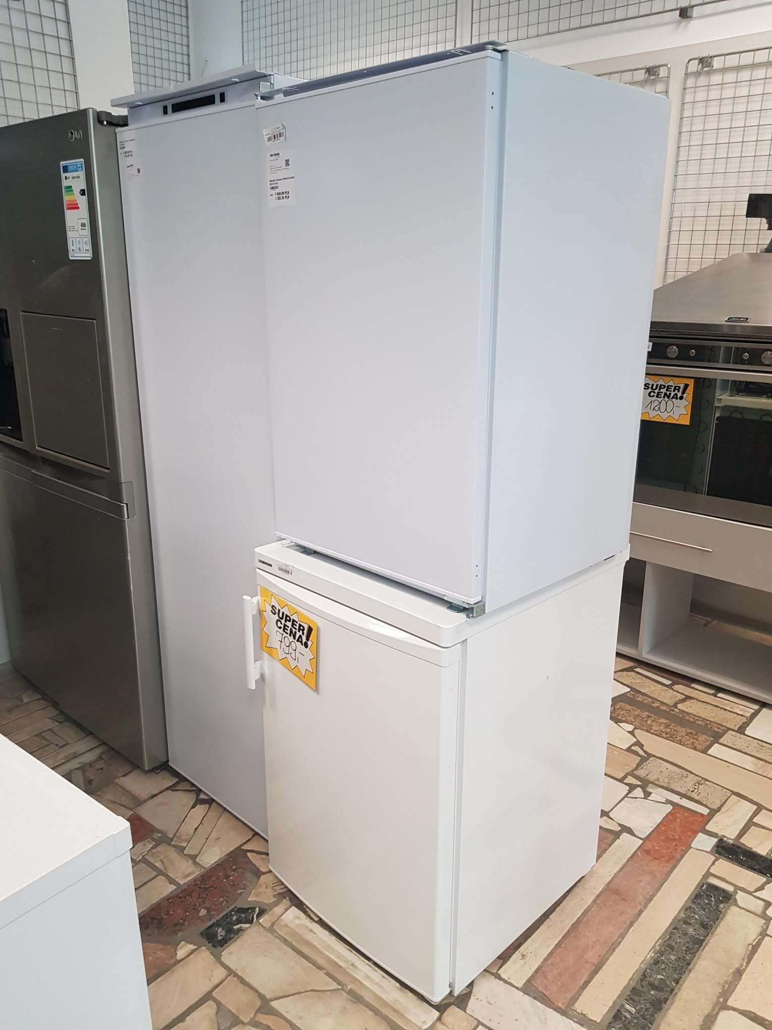 Ekspozycja w sklepie z AGD: lodówka side-by-side w kolorze srebrnym, obok biała lodówka z zamrażarką u góry oraz zamrażarka skrzyniowa postawiona pod nią. Na urządzeniach widoczne naklejki z cenami...