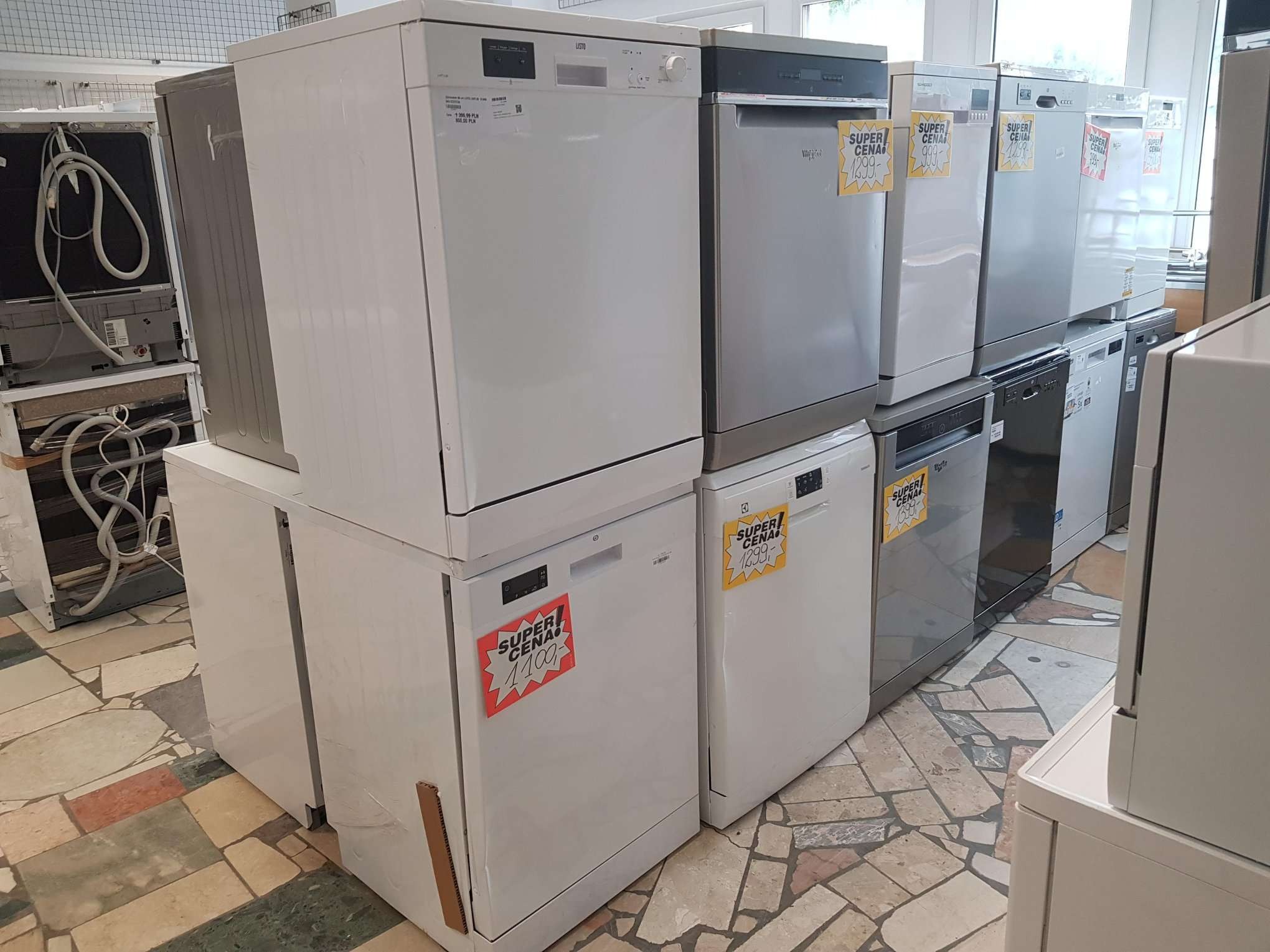 Rząd ustawionych jeden na drugim białych i szarych zmywarek różnych marek z naklejonymi żółtymi kartkami z napisem 'SUPER CENA!' i cenami w złotówkach, na tle podłogi z płytek w różnych odcieniach...