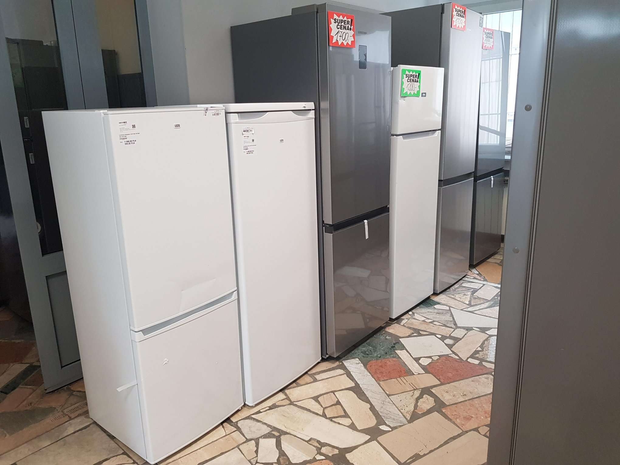 Rząd nowych lodówek w sklepie, z naklejkami promocyjnymi 'Super Cena' na niektórych modelach, ustawionych na podłodze z płytek w mozaikę.