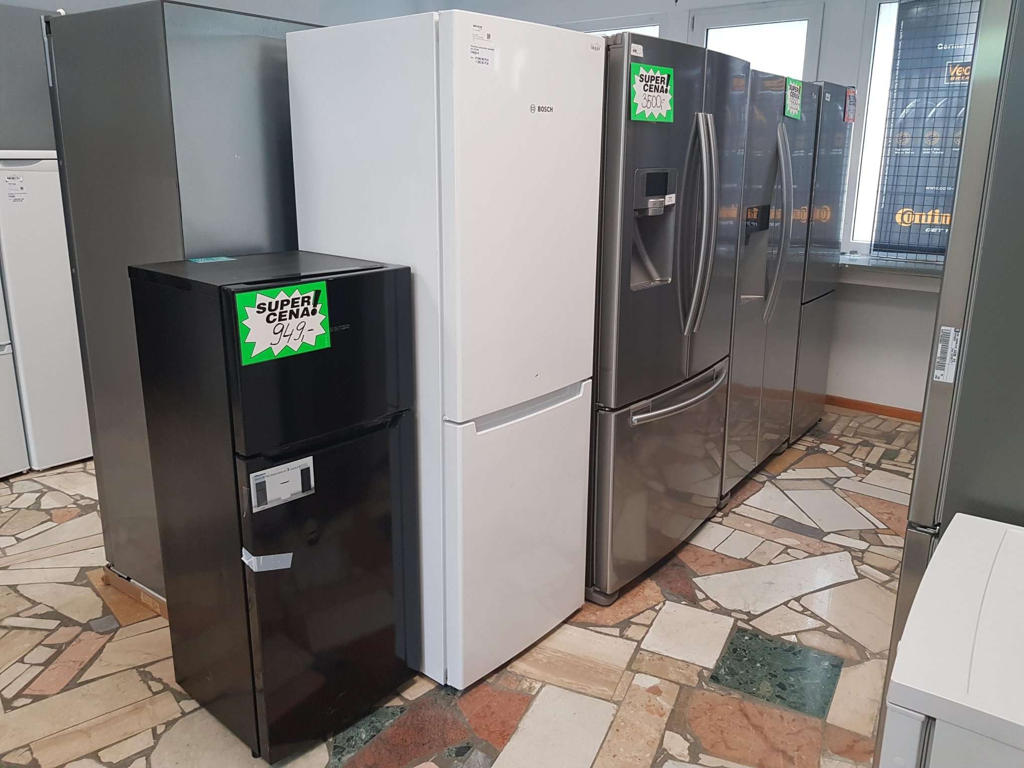 Rząd różnych modeli lodówek w sklepie, z naklejkami promocyjnymi 'Super Cena' na niektórych z nich, podłoga z mozaiki.