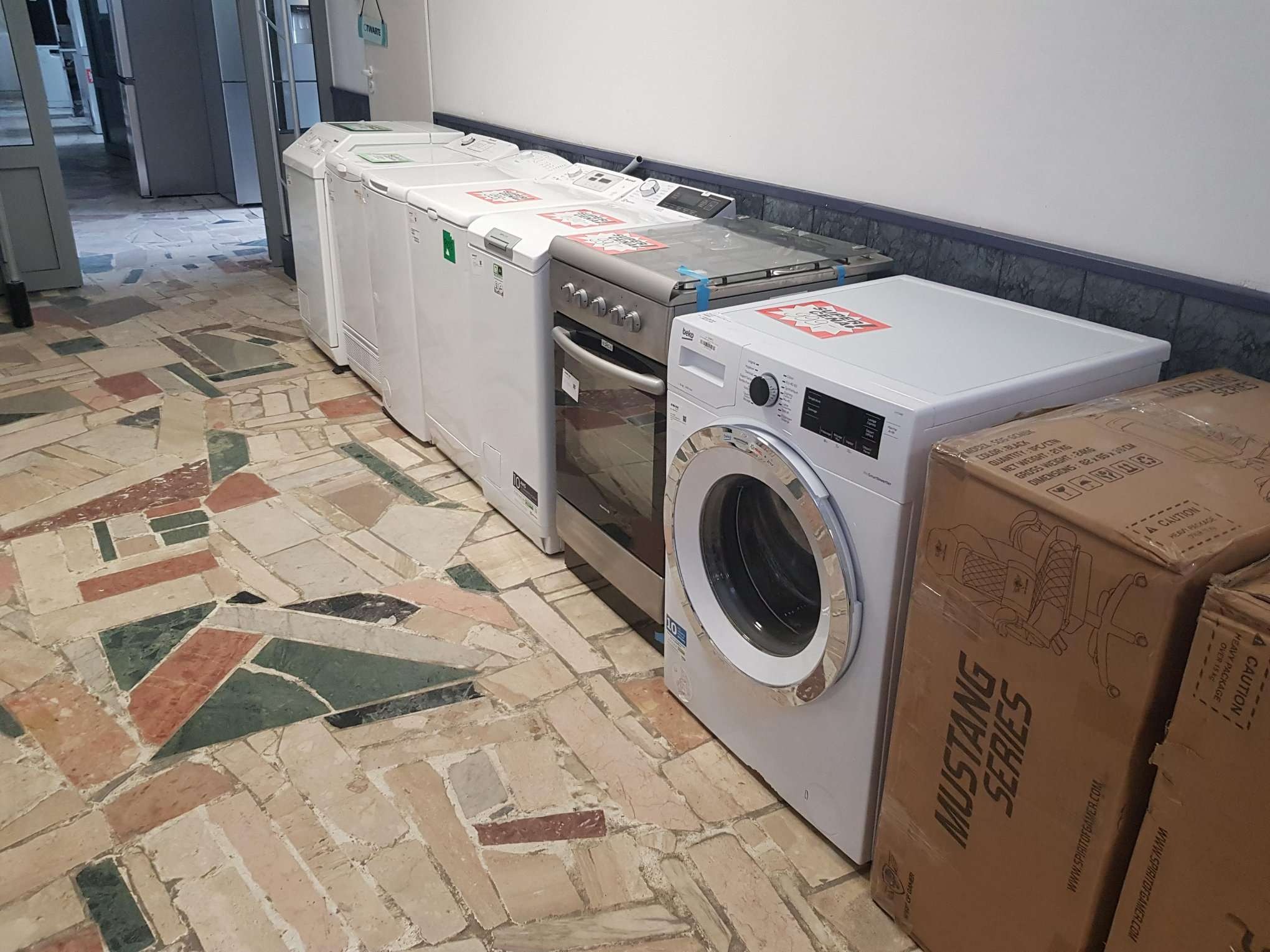 Rząd nowych, białych pralek, suszarek i piekarników ustawionych wzdłuż ściany na wzorzystej podłodze z terakoty. Część sprzętu posiada naklejki promocyjne, a obok stoi karton z nadrukiem 'Mustang...