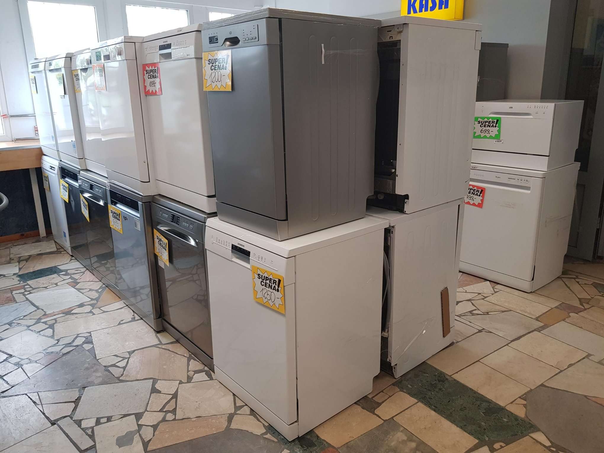 Wystawa nowych zmywarek różnych marek i kolorów z naklejkami informującymi o cenie, ustawionych na podłodze wyłożonej płytkami imitującymi kamień.