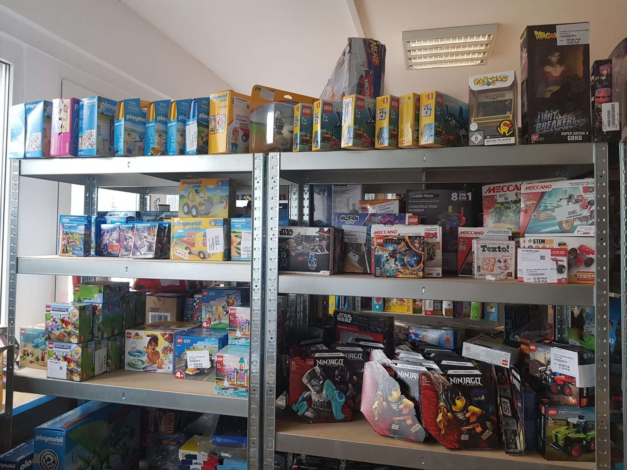 Półki magazynowe wypełnione różnorodnymi zabawkami w pudełkach: Lego, Playmobil, Nerf, Meccano, Pac-Man, Dragon Ball, Ninjago, Star Wars, Minecraft. Widoczne ceny na niektórych produktach.