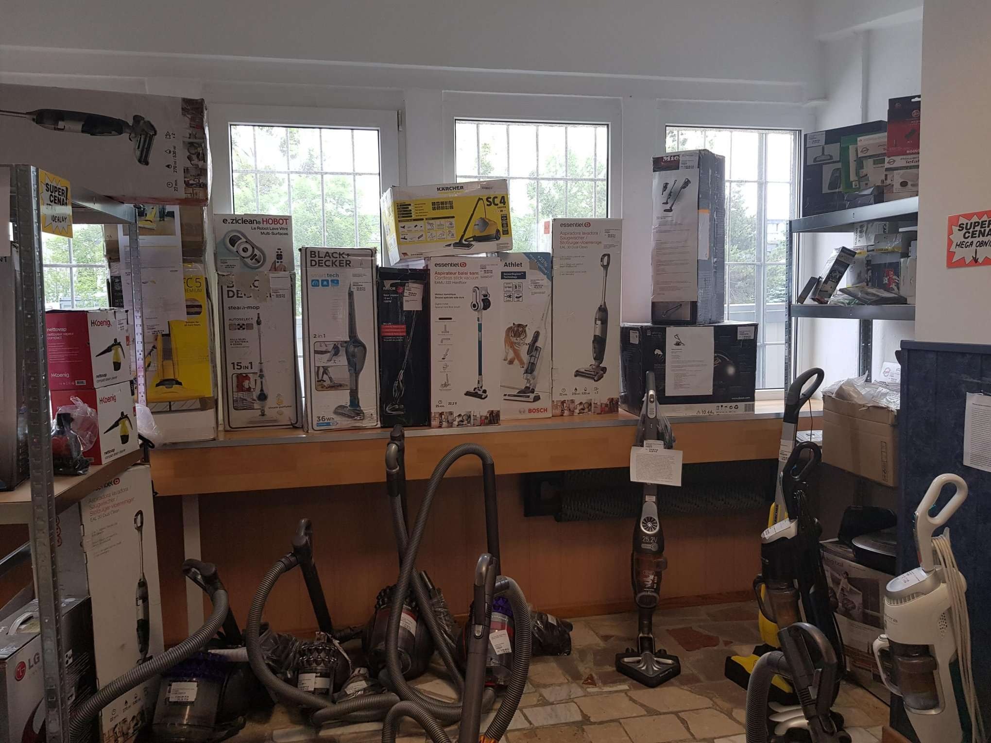 Ekspozycja różnych modeli odkurzaczy i ich opakowań w sklepie, widoczne odkurzacze pionowe, bezprzewodowe i tradycyjne, ustawione na podłodze i półkach pod oknem.