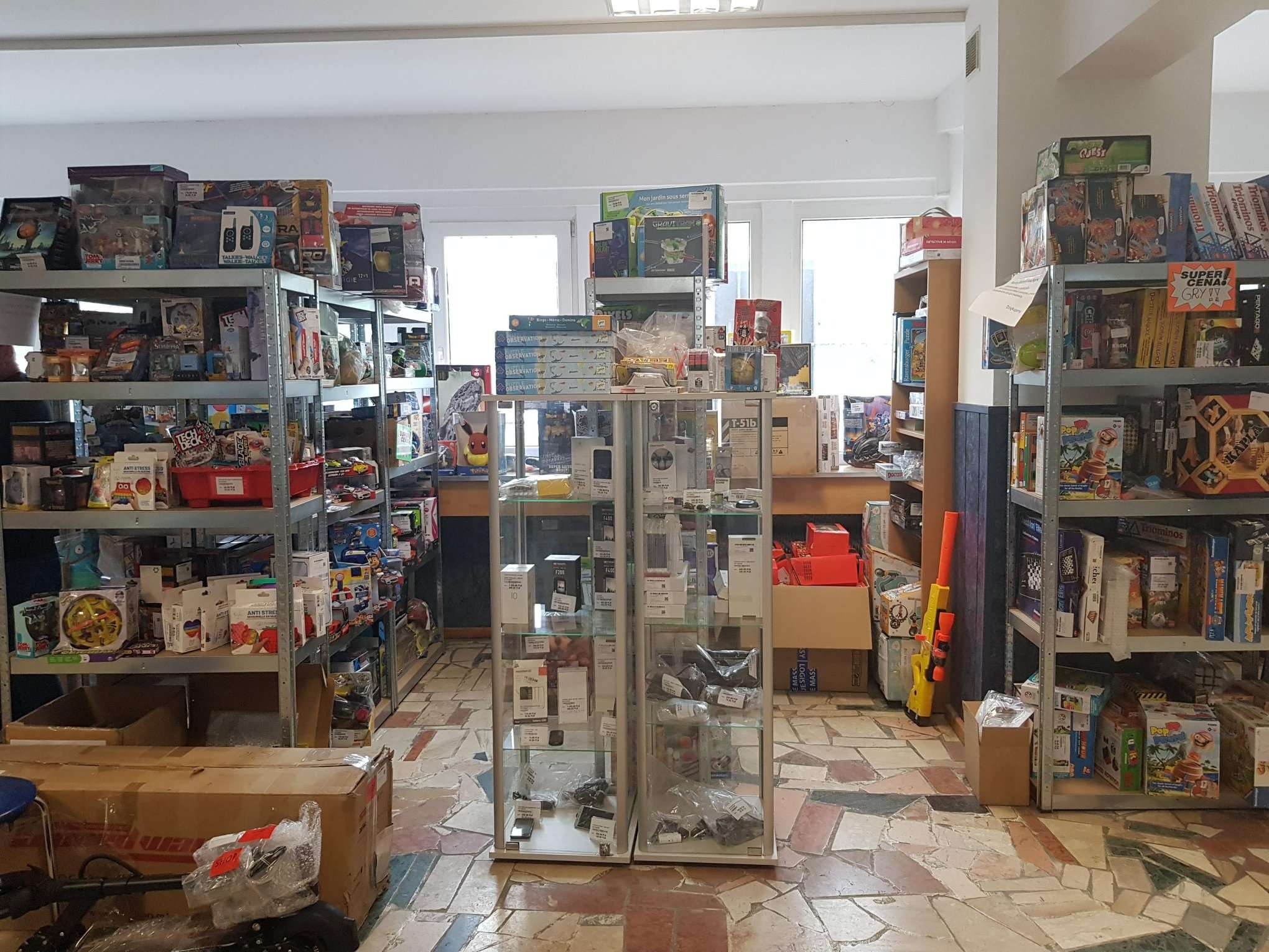 Wnętrze sklepu z zabawkami i grami planszowymi, półki metalowe z różnorodnym asortymentem, w centrum witryny szklane z elektroniką, na podłodze mozaika z płytek.