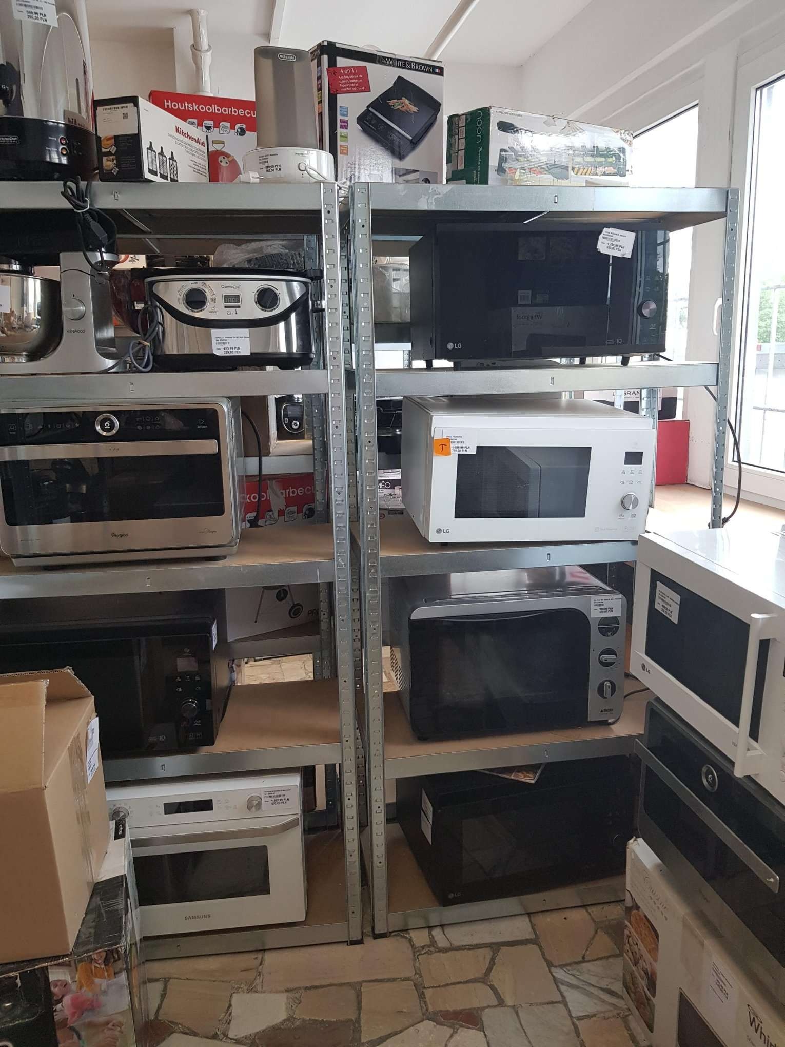 Półki magazynowe wypełnione różnymi modelami kuchenek mikrofalowych i robotów kuchennych, w tym marki LG, Samsung, Kenwood i Whirlpool, widoczne etykiety cenowe.