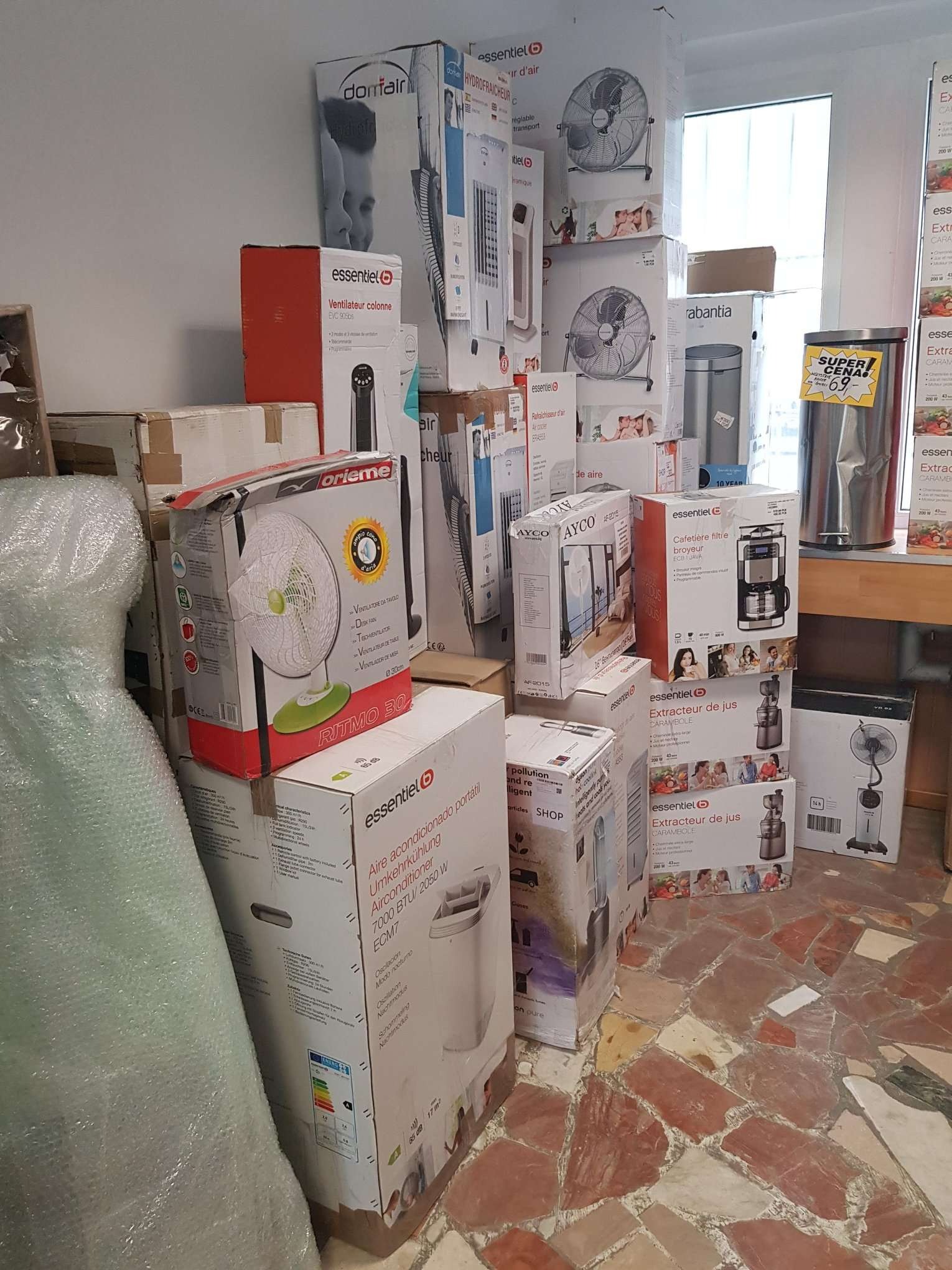Stos pudełek z wentylatorami, klimatyzatorami przenośnymi, wyciskarkami do soków, ekspresami do kawy i koszami na śmieci różnych marek, ustawionych na podłodze w sklepie lub magazynie.