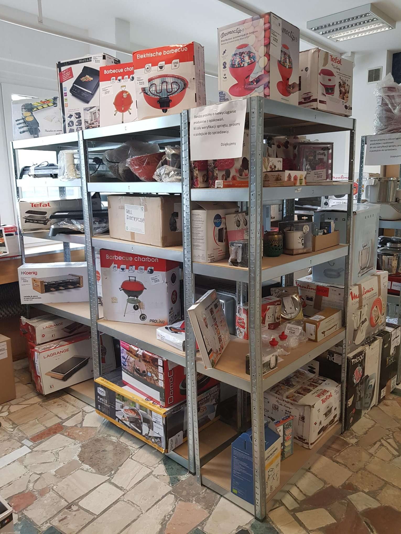Metalowy regał magazynowy wypełniony kartonami z urządzeniami AGD, głównie grillami elektrycznymi, raclette i dystrybutorami słodyczy, w pomieszczeniu z mozaikową podłogą.