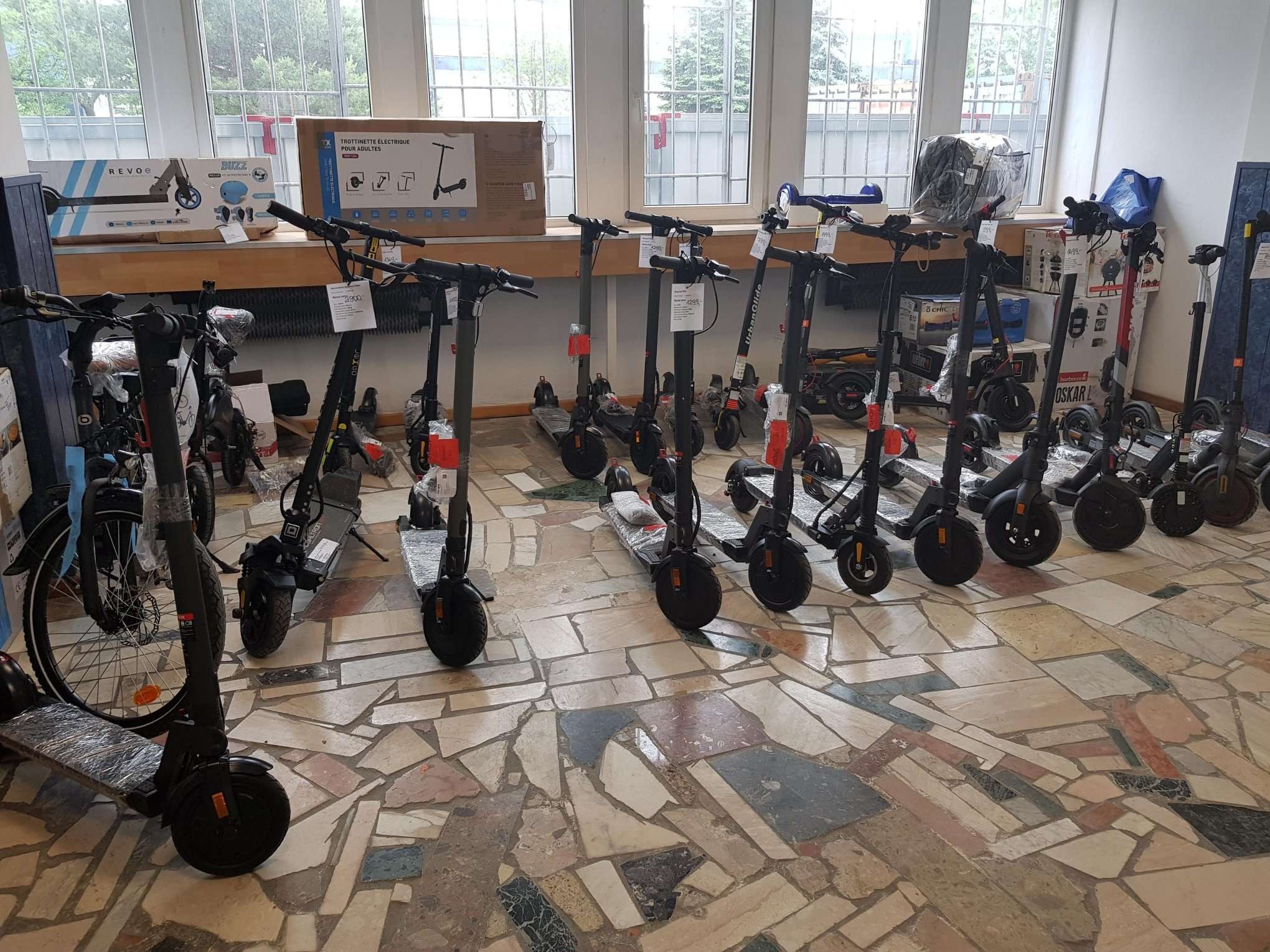 Wystawa skuterów elektrycznych różnych marek i modeli w salonie sprzedaży, widoczne ceny na etykietach, opakowania i akcesoria w tle, posadzka z mozaiki.