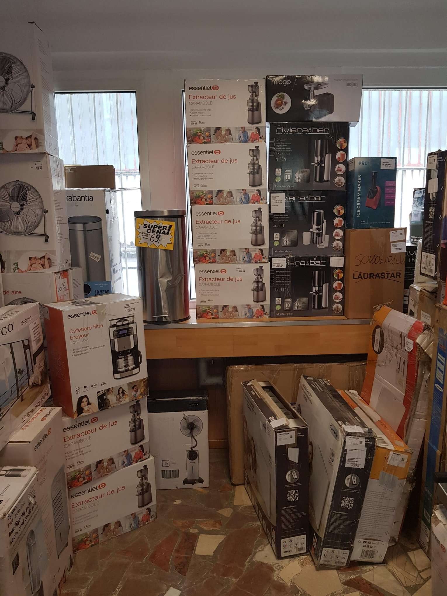 Stos pudełek z urządzeniami AGD, w tym wyciskarki do soków, ekspresy do kawy, wentylatory i roboty kuchenne, ustawione w pomieszczeniu przypominającym magazyn lub sklep.