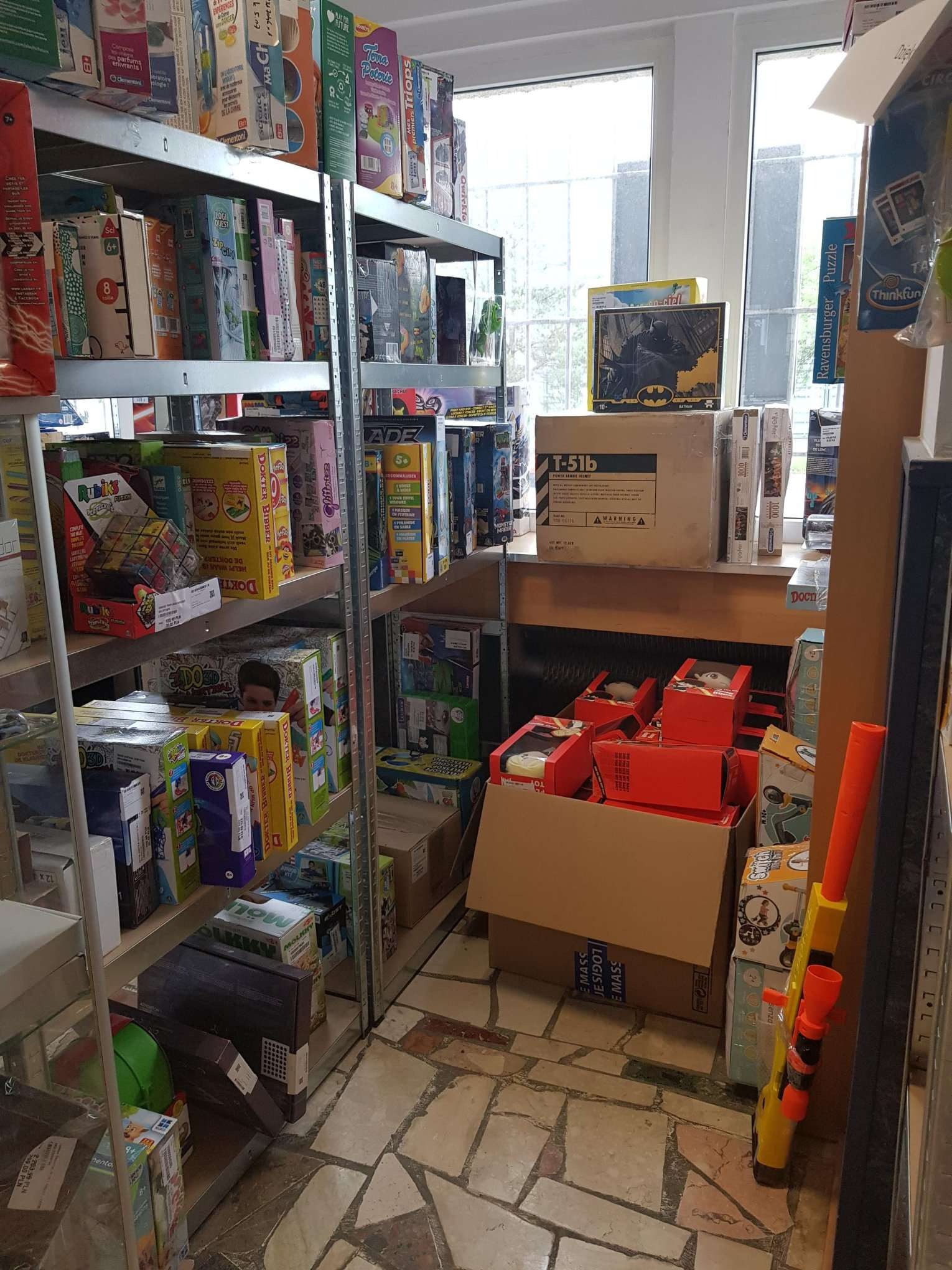 Półki metalowe wypełnione różnorodnymi grami planszowymi i zabawkami, widoczne okna w tle, na podłodze kartony z zabawkami, obok żółto-pomarańczowa zabawka przypominająca karabin.