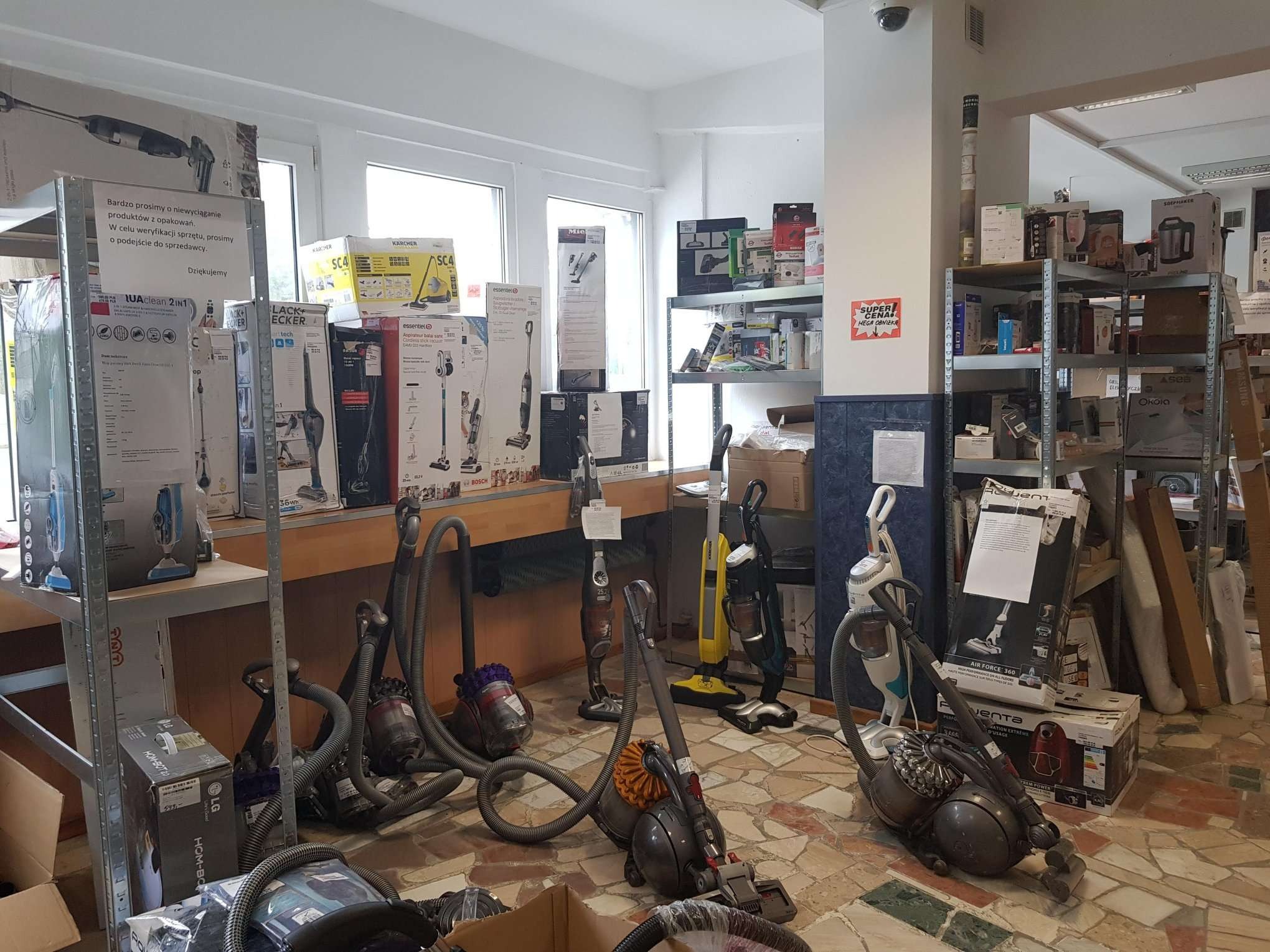 Wnętrze sklepu z ekspozycją odkurzaczy różnych marek, w tym Dyson, Karcher i Black+Decker, zarówno w pudełkach, jak i rozpakowanych modelach demonstracyjnych.