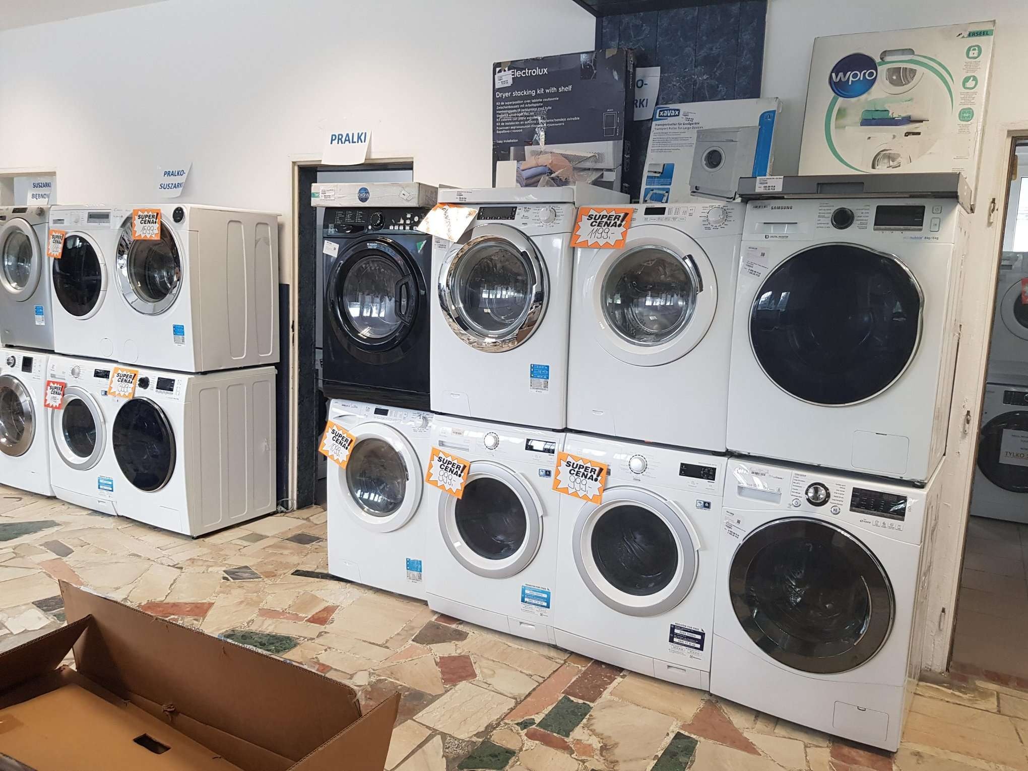 Ekspozycja nowych pralek i pralko-suszarek różnych marek, w tym czarnej pralki, z cenami promocyjnymi w sklepie AGD w Łodzi. Na podłodze widoczny karton.