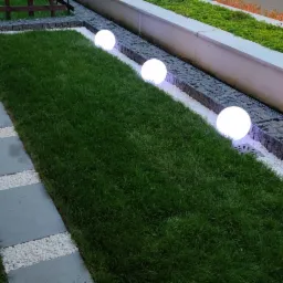 Świeżo założony trawnik z rolki, oświetlony kulistymi lampami, z ścieżką z płyt chodnikowych i białego grysu, oraz gabionami wzdłuż krawędzi.