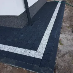 Ułożona, ciemnoszara kostka brukowa z białym pasem dekoracyjnym obok ściany budynku z rurą spustową i kranem.