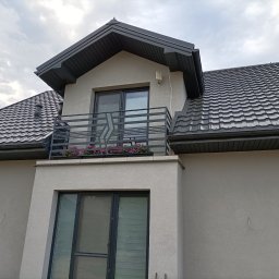 WELDEC Mobilne usługi spawalniczo-ślusarskie - Nowoczesny dom z balkonem, na którym zamontowano czarną, metalową balustradę. Na balkonie widoczne kwiaty w donicach. Dach pokryty grafitową blachodachówką.