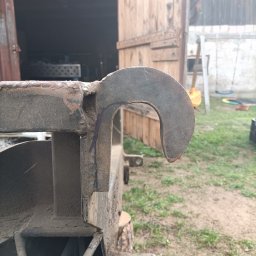 WELDEC Mobilne usługi spawalniczo-ślusarskie - Metalowy hak spawalniczy z wyraźnymi śladami spawania, uchwycony w zbliżeniu na tle otwartego warsztatu i fragmentu podwórza z trawą.