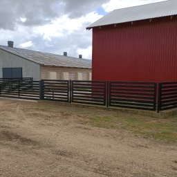 WELDEC Mobilne usługi spawalniczo-ślusarskie - Nowoczesna, pozioma, czarna balustrada posesji na tle czerwonego budynku gospodarczego i szarego magazynu pod pochmurnym niebem. Detal: betonowa podstawa.