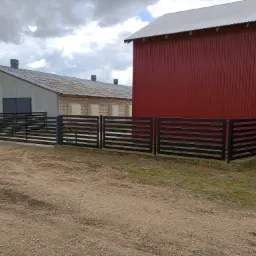 Nowoczesna, pozioma, czarna balustrada posesji na tle czerwonego budynku gospodarczego i szarego magazynu pod pochmurnym niebem. Detal: betonowa podstawa.