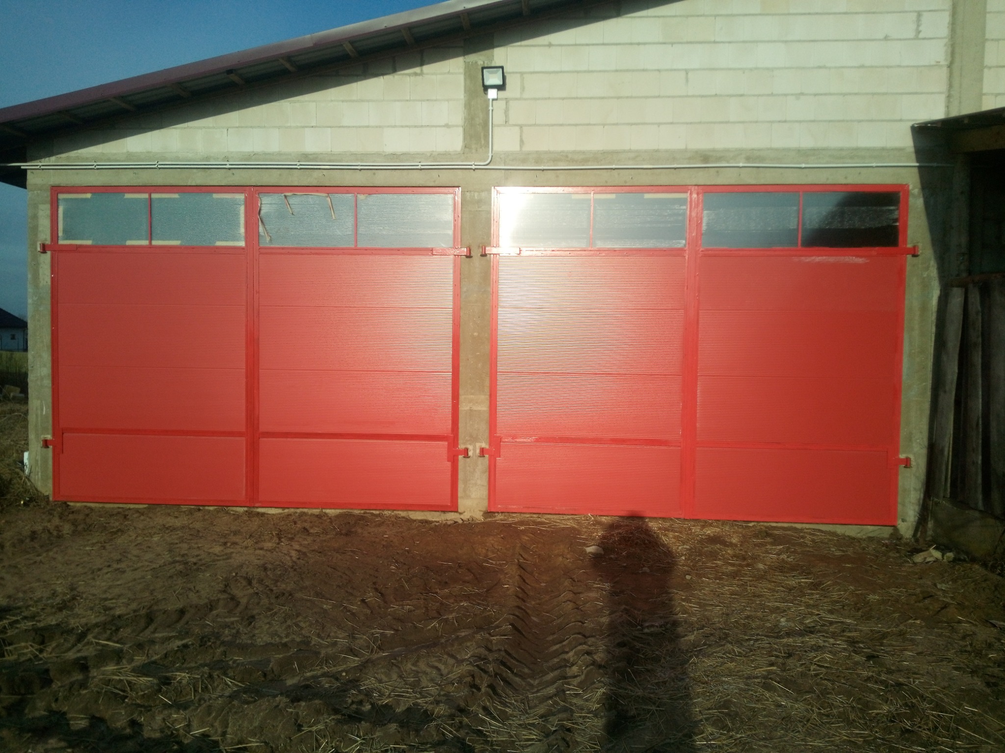 Czerwona, dwuskrzydłowa brama z oknami w hali magazynowej, z widocznym oświetleniem zewnętrznym i śladami opon na ziemi. Brama z blachy trapezowej.