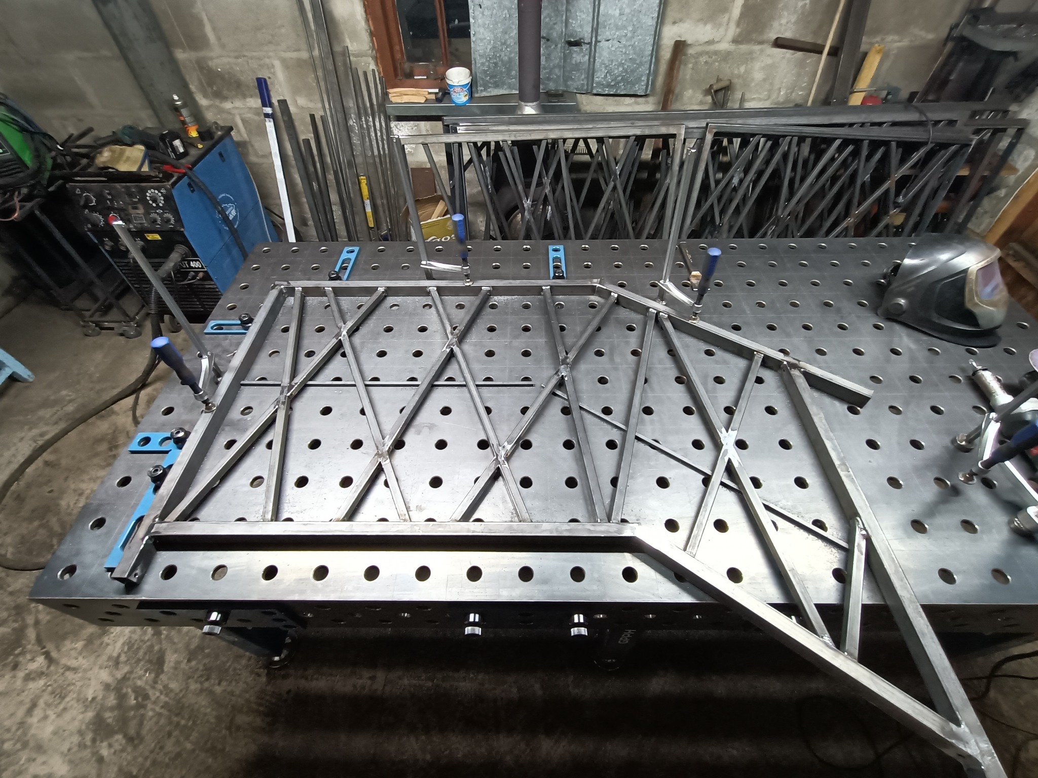 Konstrukcja metalowa z ukośnymi wzmocnieniami, mocowana klamrami na perforowanym stole spawalniczym. W tle widoczne narzędzia i elementy konstrukcji.