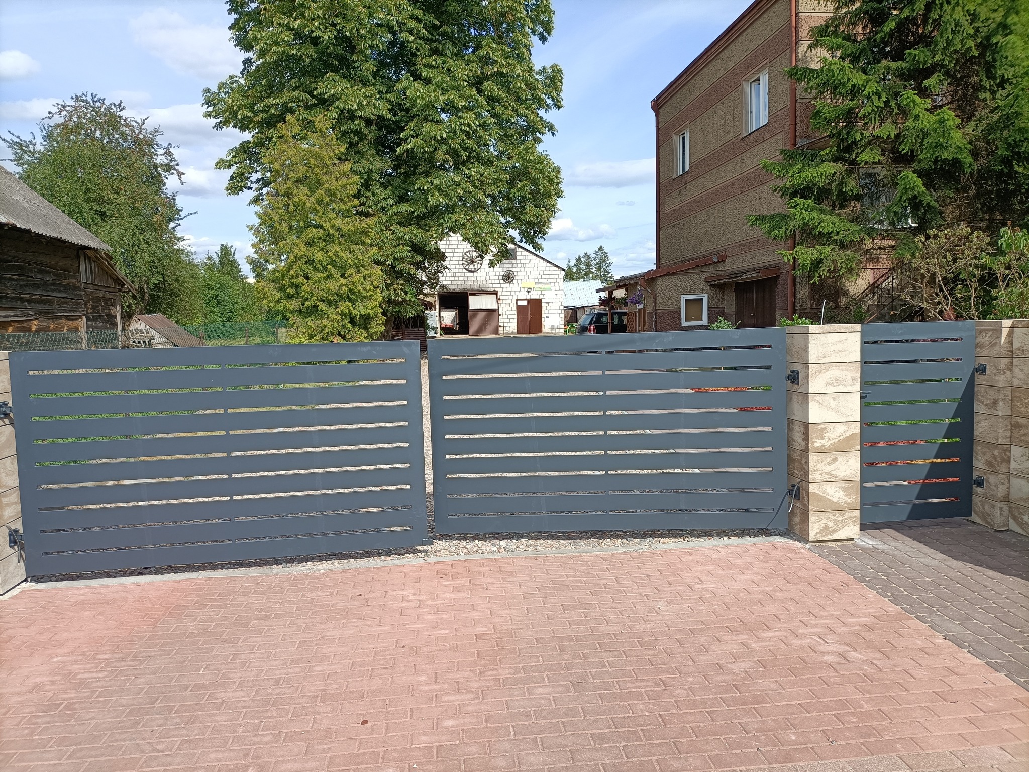 Nowoczesna, pozioma brama posesyjna w kolorze grafitowym, z jasnymi, kamiennymi słupkami w Suchowoli. Brama otwarta, widoczny fragment posesji.