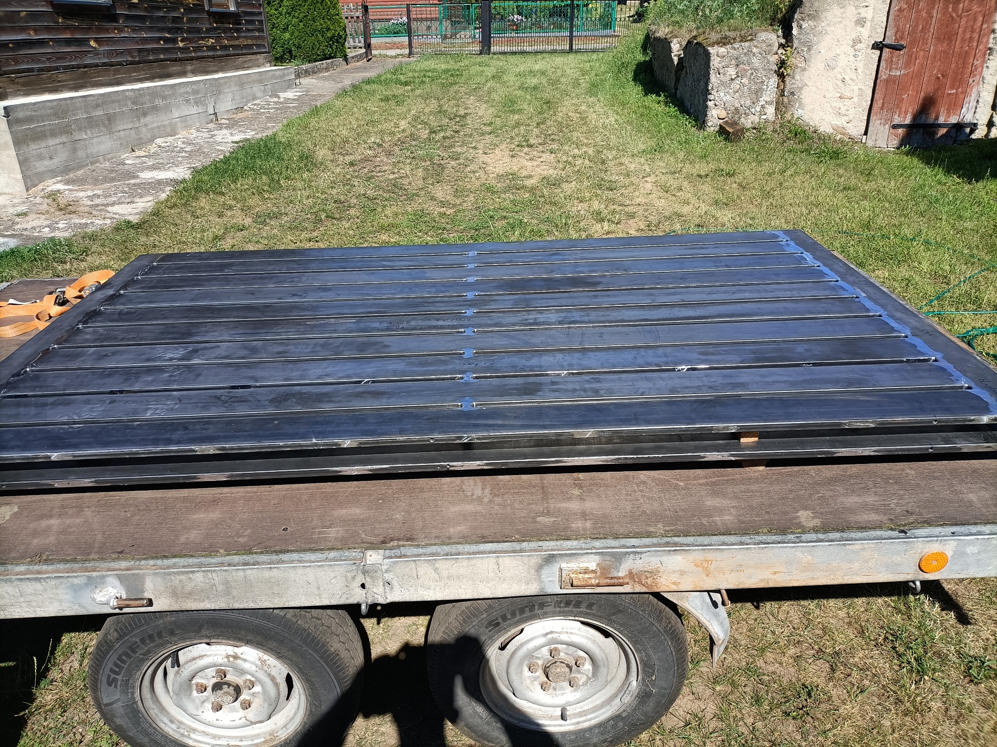 Ciemna, spawana konstrukcja stalowa z prętów, zabezpieczona taśmą, leży na przyczepce samochodowej na tle trawy i budynków. Widoczne koła przyczepki.