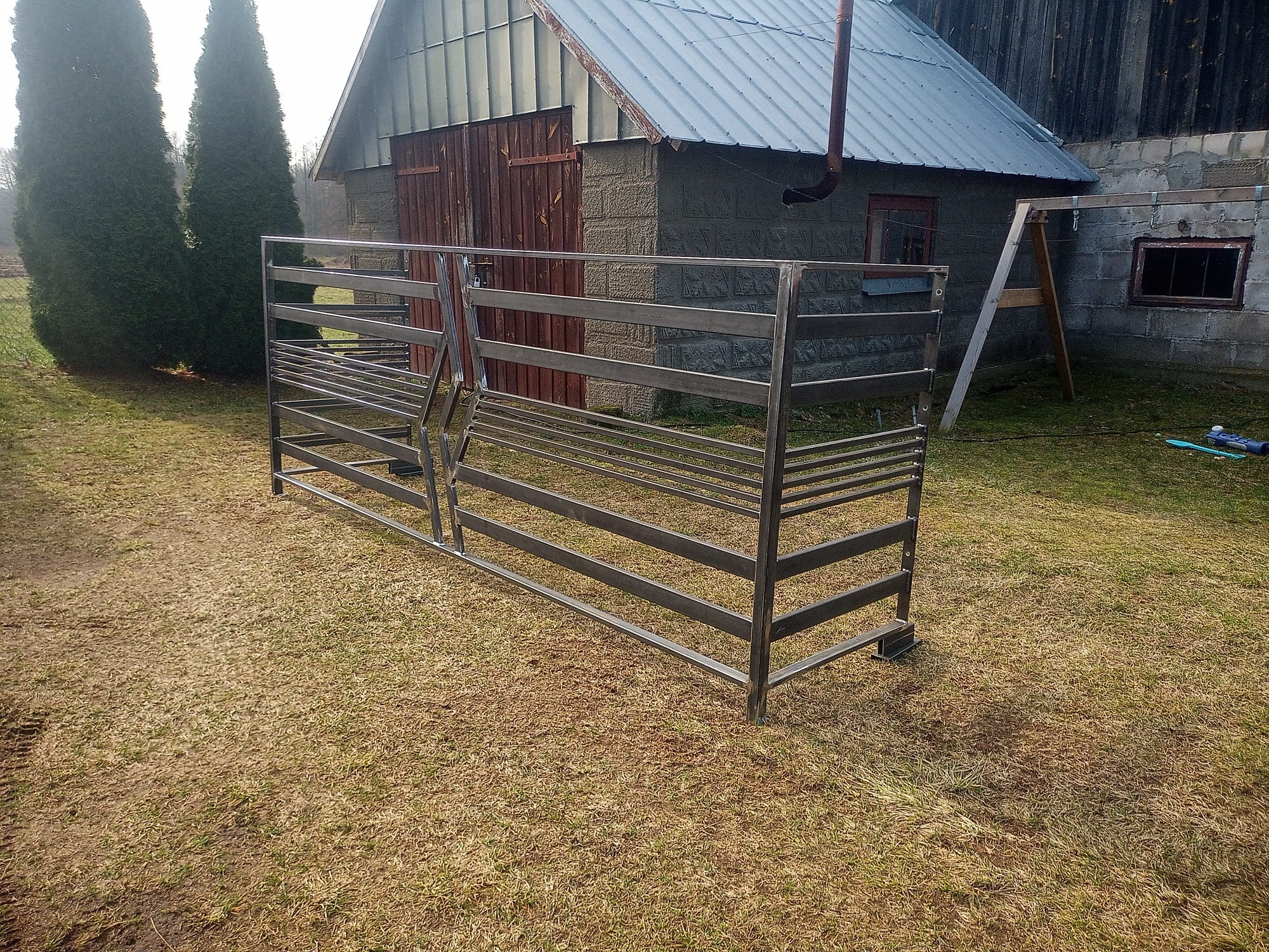 Metalowa konstrukcja bramy/kojca na trawiastym terenie, z budynkiem gospodarczym w tle. Konstrukcja spawana z profili, surowa, bez wykończenia. Widok z boku, lekko z dołu.