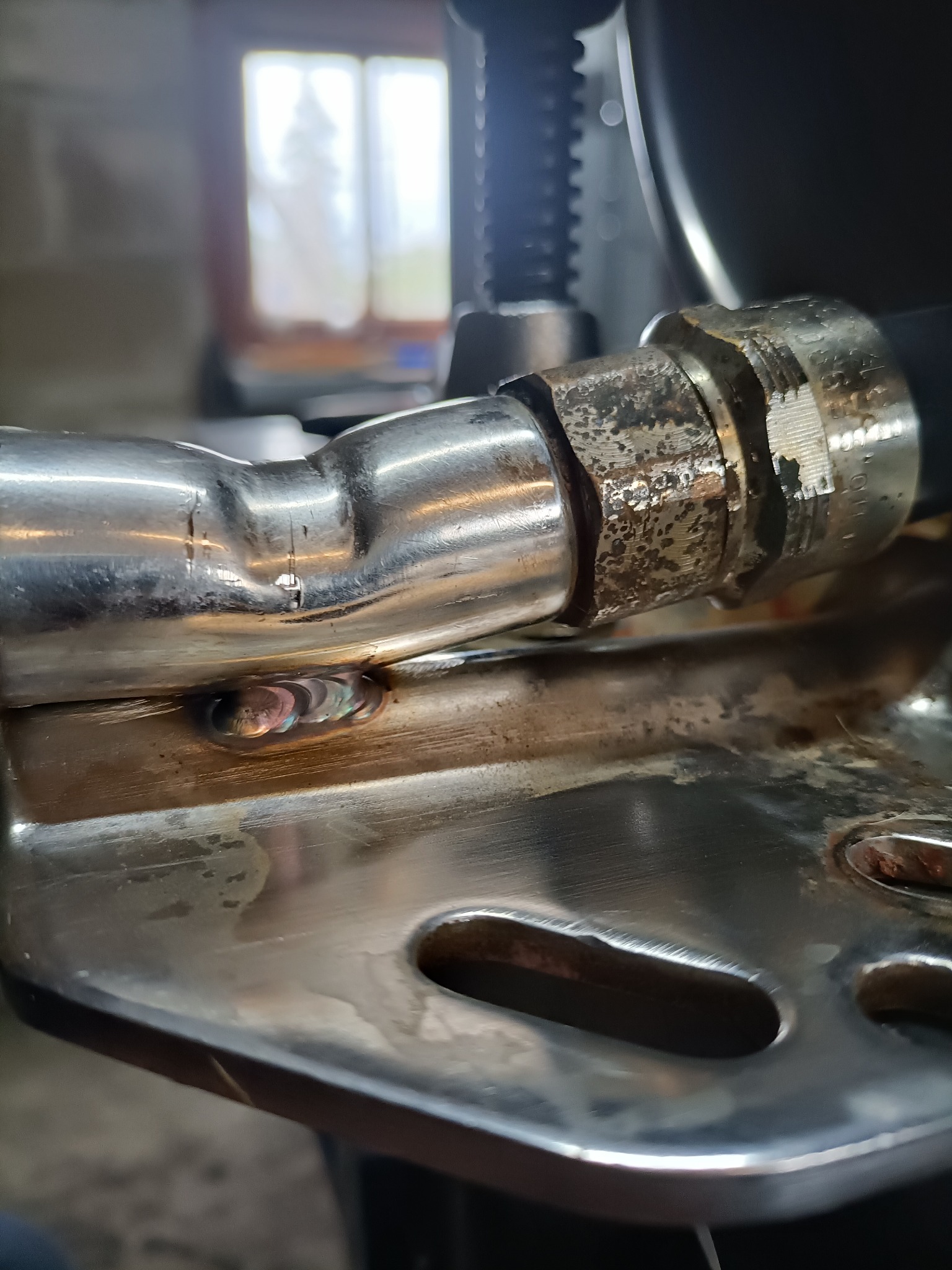 Szlifowana spoina na metalowej rurze w zbliżeniu, widoczne tęczowe przebarwienia po spawaniu. Tło rozmyte, z widocznym oknem.