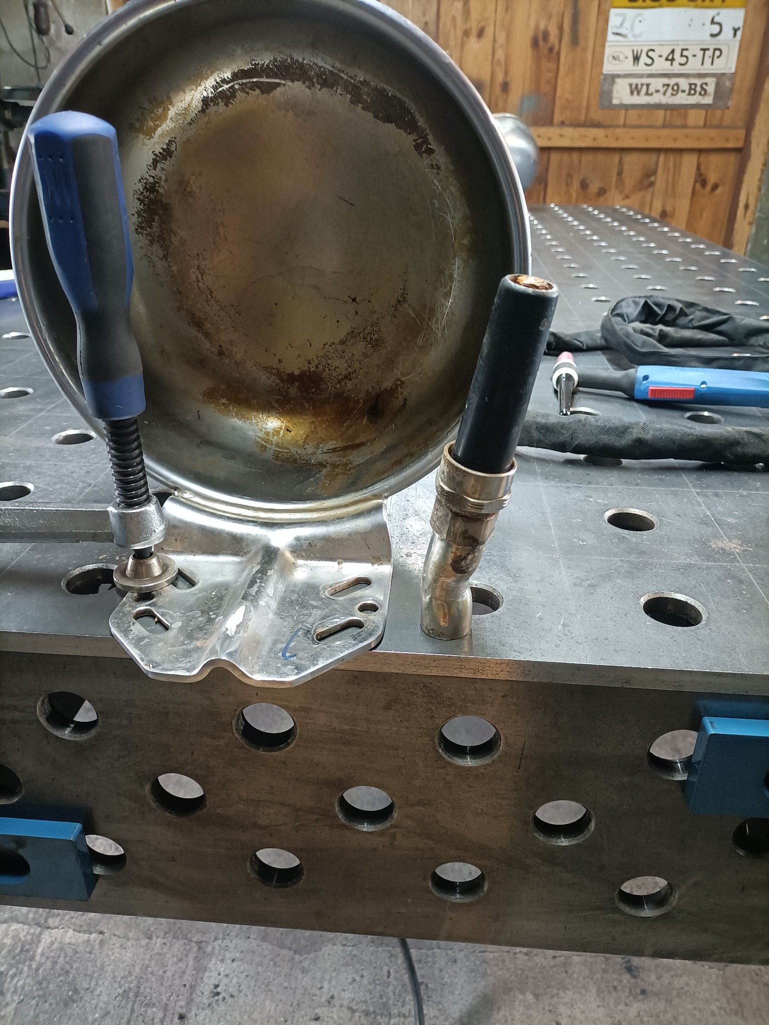 Metalowy element w uchwycie na perforowanym stole spawalniczym, widoczny uchwyt spawalniczy i narzędzia. Przygotowanie do spawania elementu w warsztacie.