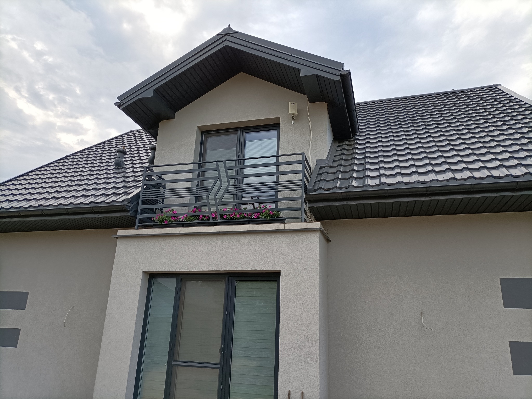 Nowoczesny dom z balkonem, na którym zamontowano czarną, metalową balustradę. Na balkonie widoczne kwiaty w donicach. Dach pokryty grafitową blachodachówką.