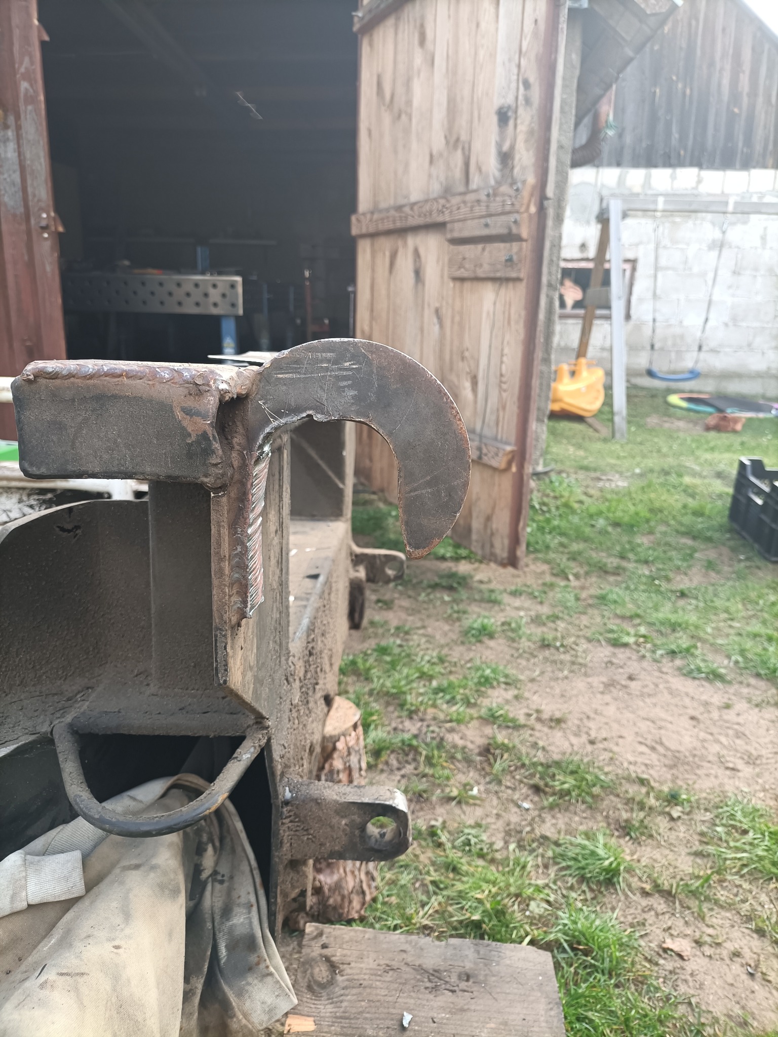 Metalowy element z widocznymi spawami, przypominający hak, przymocowany do konstrukcji. W tle drewniane drzwi stodoły i trawa.