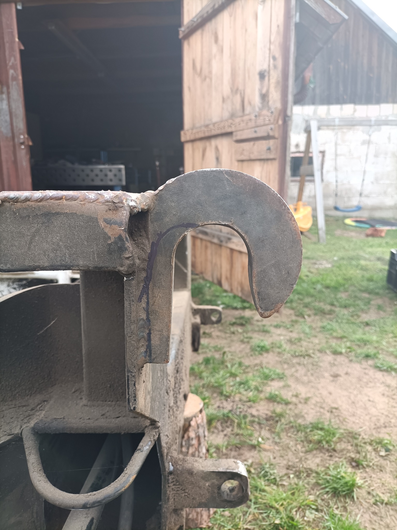 Metalowy hak spawalniczy z wyraźnymi śladami spawania, uchwycony w zbliżeniu na tle otwartego warsztatu i fragmentu podwórza z trawą.