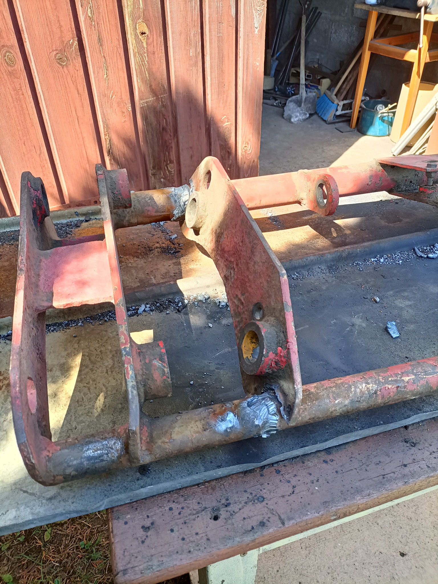 Częściowo odnowiona, spawana konstrukcja metalowa pomalowana na czerwono, z widocznymi śladami spawania, leżąca na drewnianym podłożu, w warsztatowym otoczeniu.