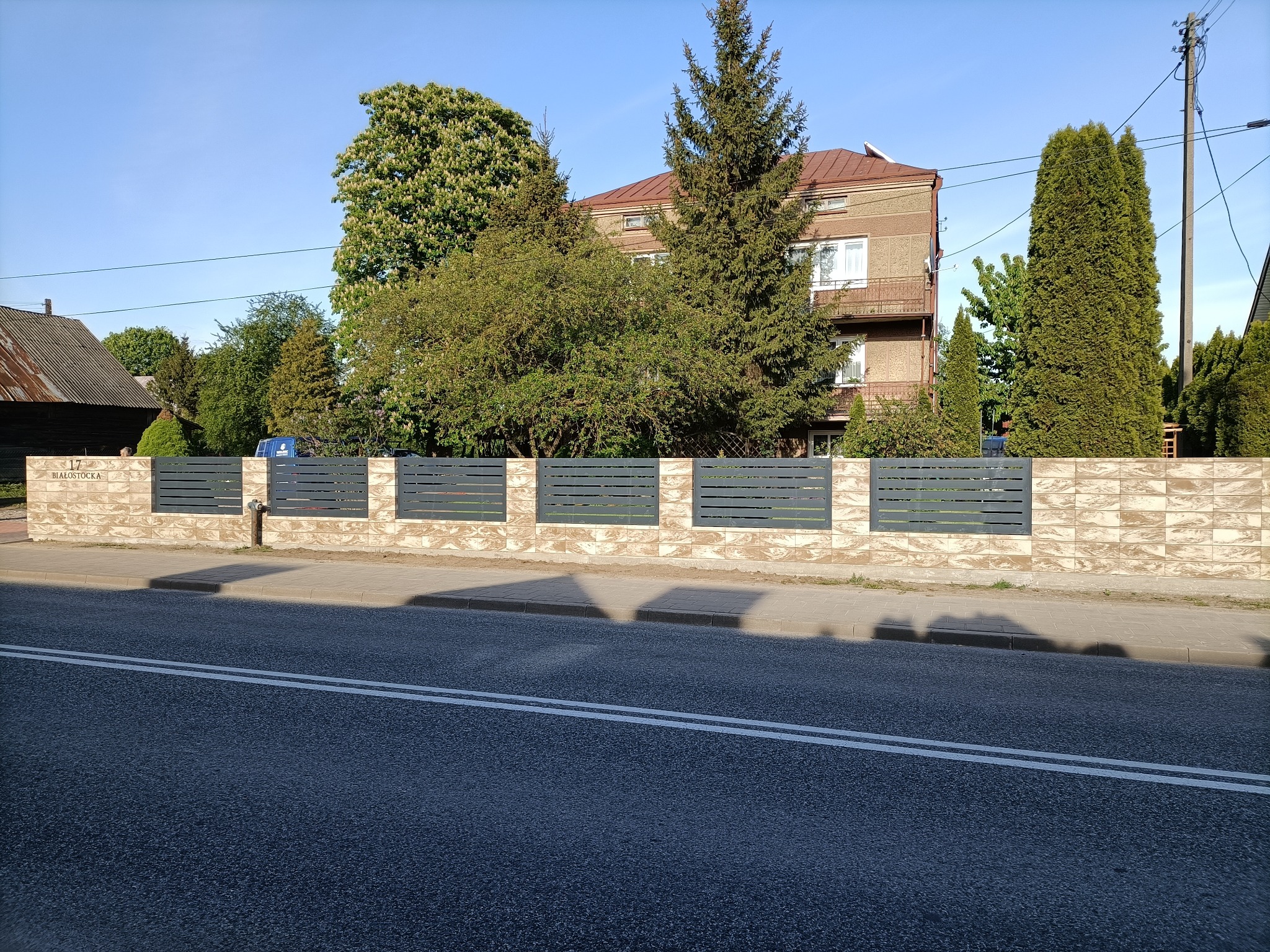 Nowoczesne, poziome, szare przęsła ogrodzeniowe zamontowane na niskim murku z beżowych płytek, na tle dwupiętrowego budynku w otoczeniu zieleni.