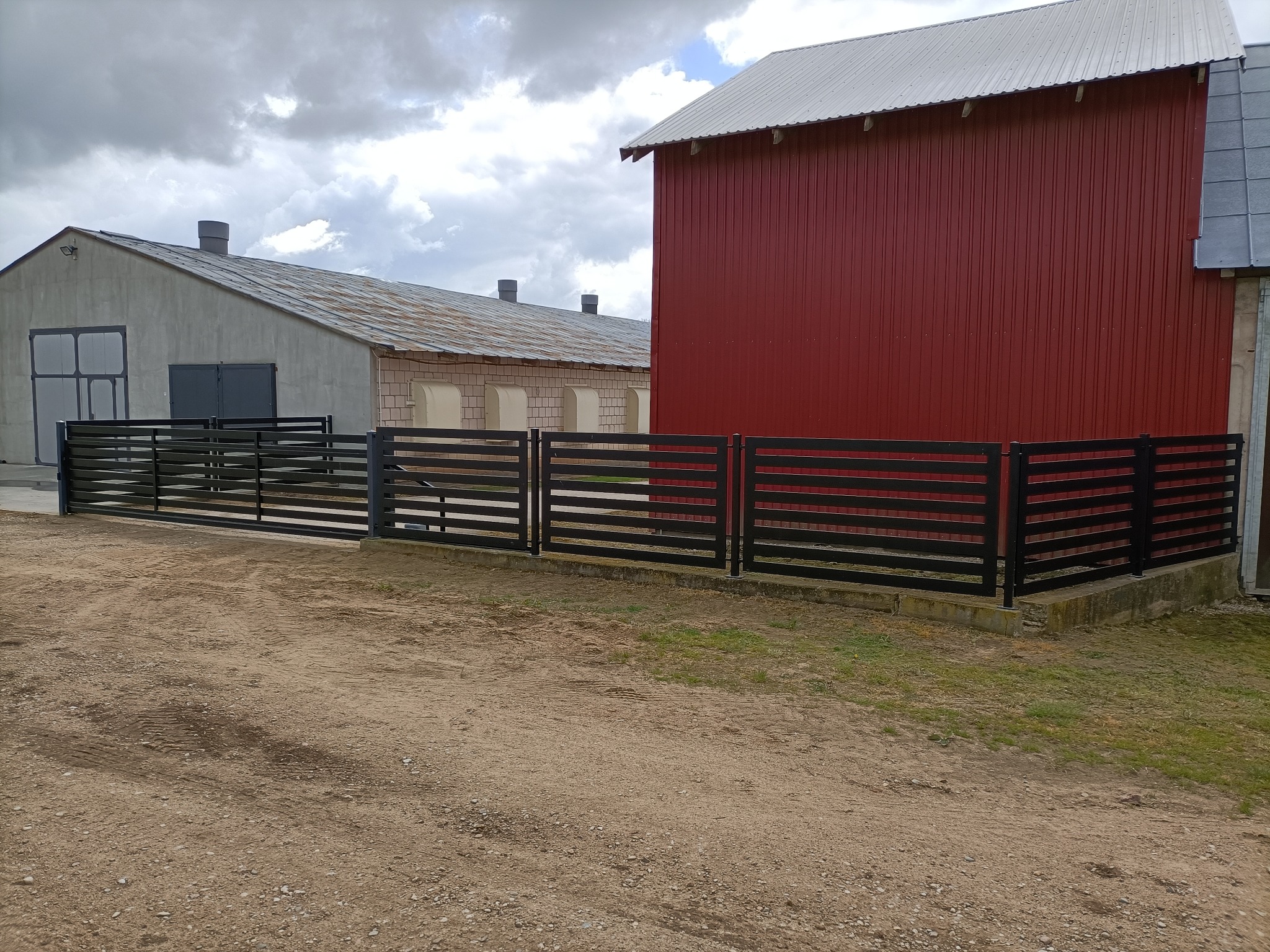 Nowoczesna, pozioma, czarna balustrada posesji na tle czerwonego budynku gospodarczego i szarego magazynu pod pochmurnym niebem. Detal: betonowa podstawa.