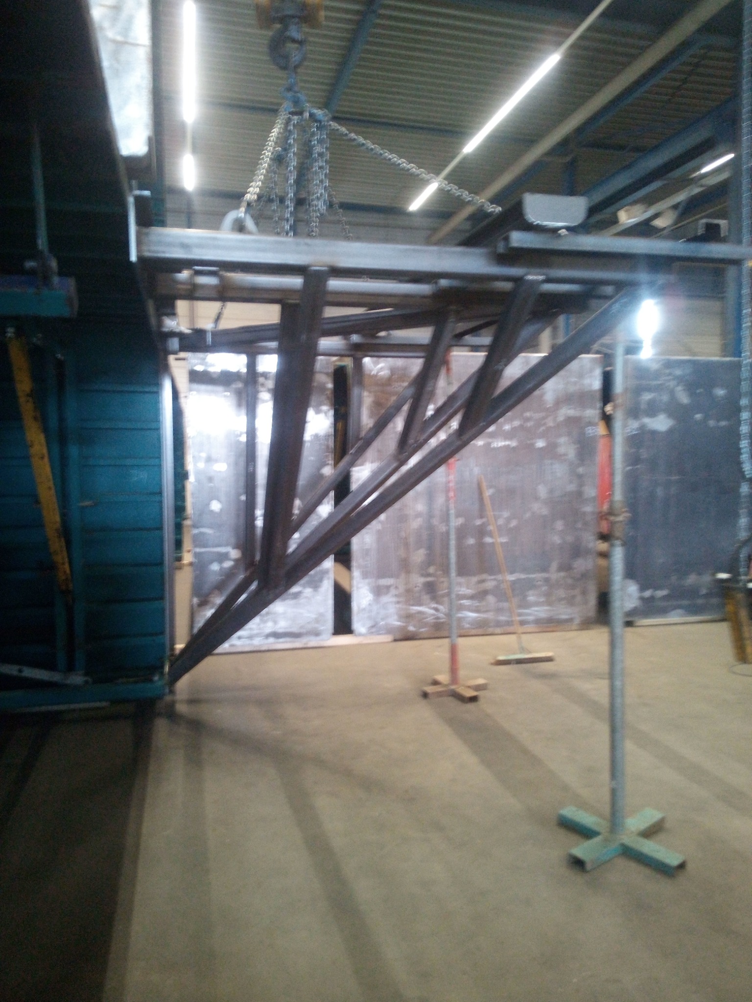 Metalowa konstrukcja spawana podwieszona na łańcuchach, w hali produkcyjnej, widoczne podpory i narzędzia.