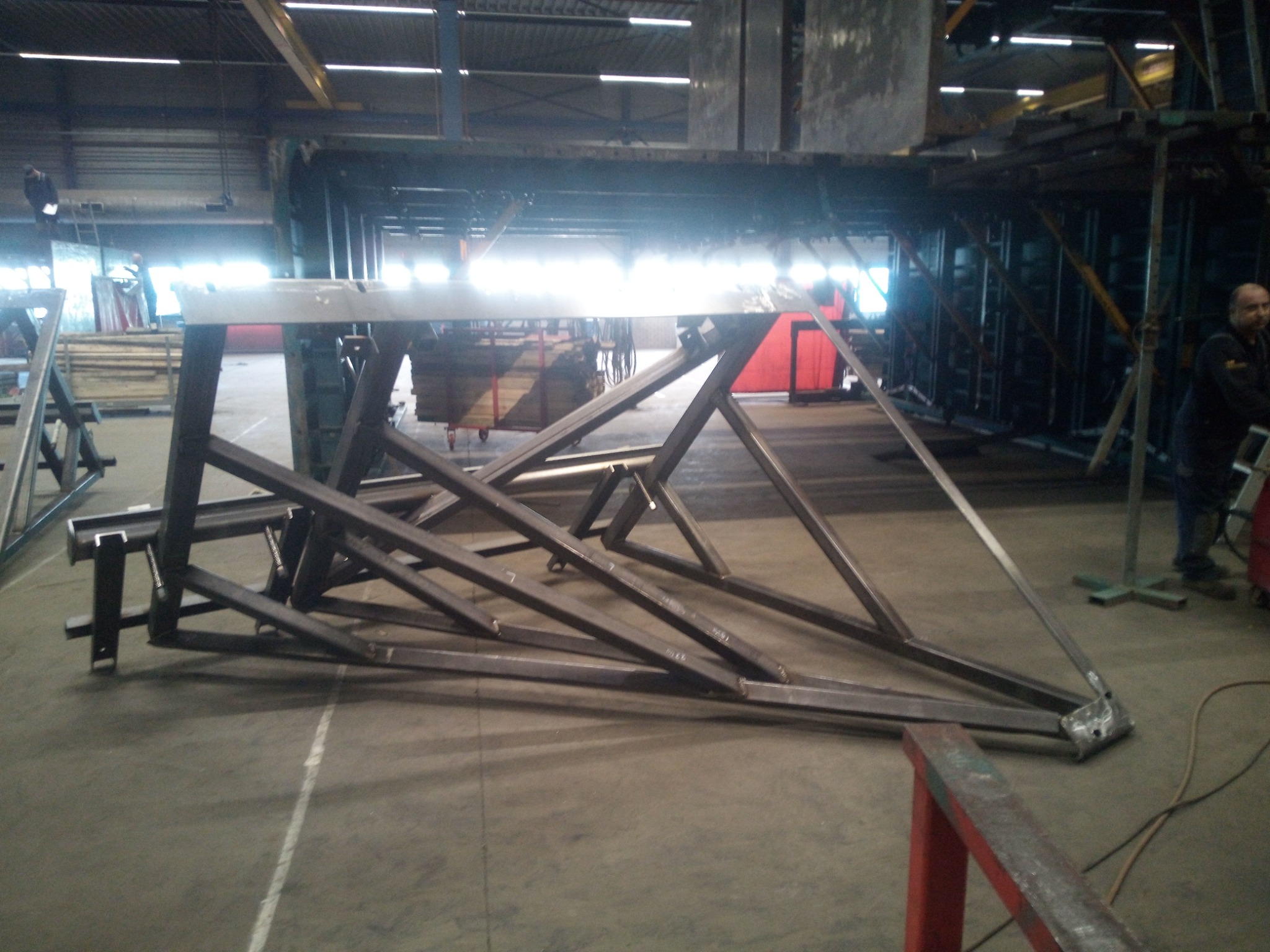 Metalowa konstrukcja szkieletowa z profili stalowych w hali produkcyjnej, widoczne spawy i elementy mocujące, w tle inne konstrukcje i osoba w roboczym ubraniu.