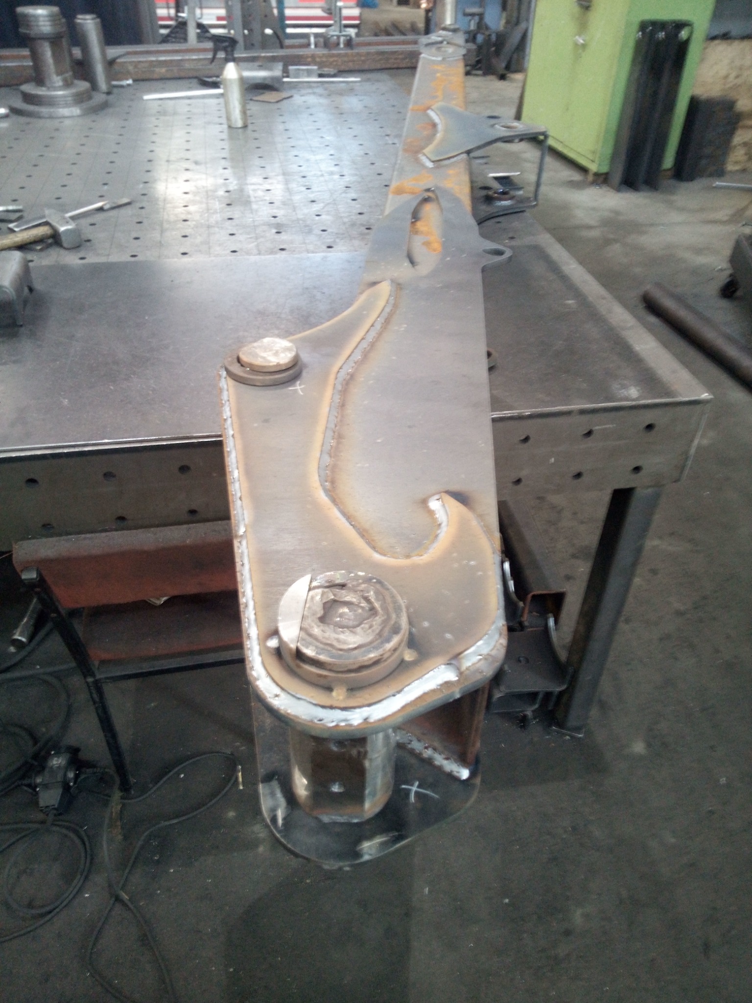 Metalowy element konstrukcyjny ze śladami spawania leżący na stole spawalniczym z widocznymi narzędziami i wyposażeniem warsztatu w tle.