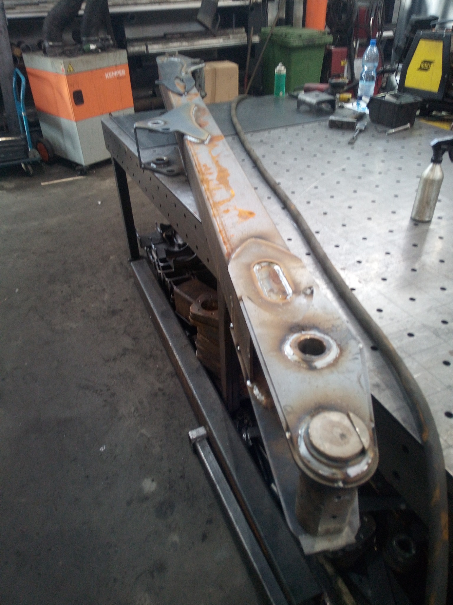 Metalowa konstrukcja spawana, leżąca na stole warsztatowym, widoczne spawy i ślady obróbki, w tle sprzęt warsztatowy i podłoga z betonu.