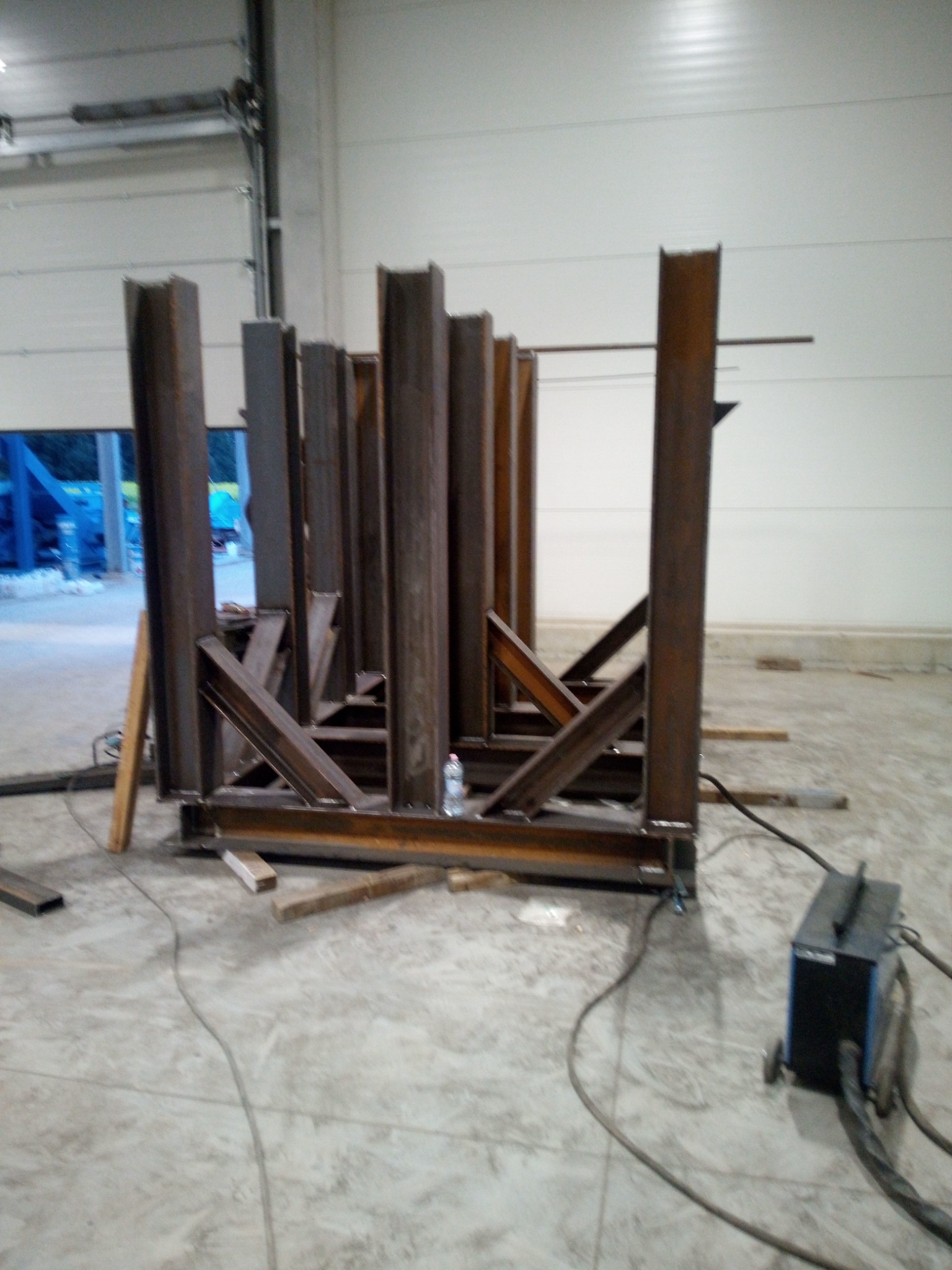 Konstrukcja stalowa z profili zamkniętych w hali, widoczny spawarka i otwarte drzwi hali w tle.