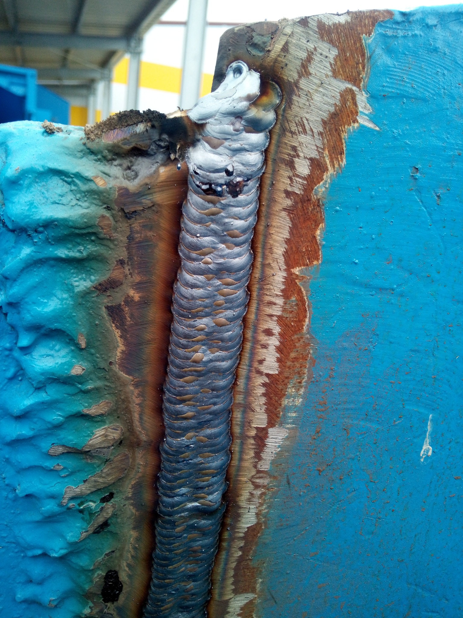 Świeżo wykonane spoiny na grubym, pomalowanym na niebiesko elemencie konstrukcyjnym, widoczne ślady obróbki cieplnej i odpryski farby wzdłuż krawędzi spoiny.