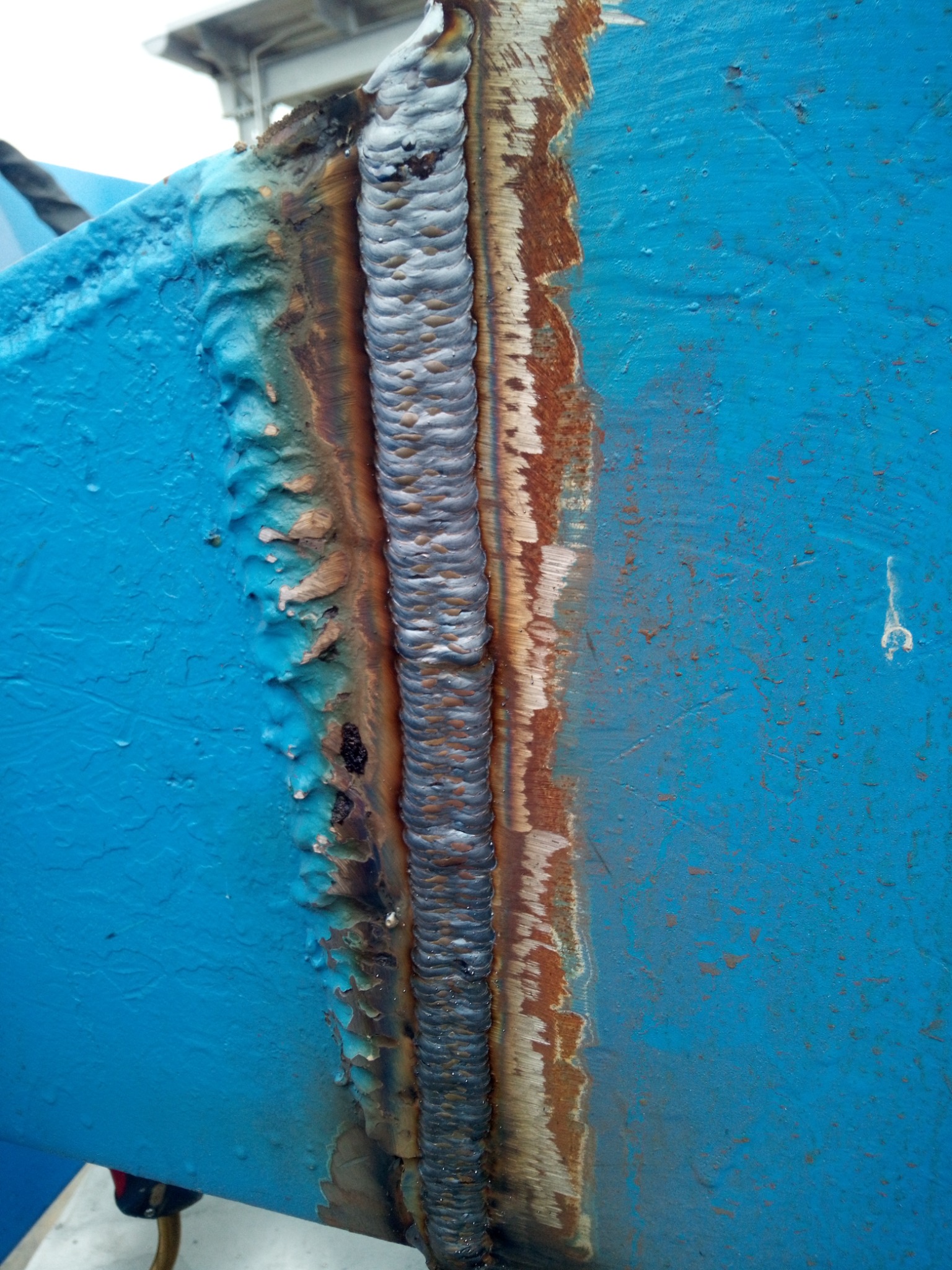 Świeżo wykonany spaw na konstrukcji pomalowanej na niebiesko, widoczne ślady odprysków farby i rdzy wzdłuż spoiny.