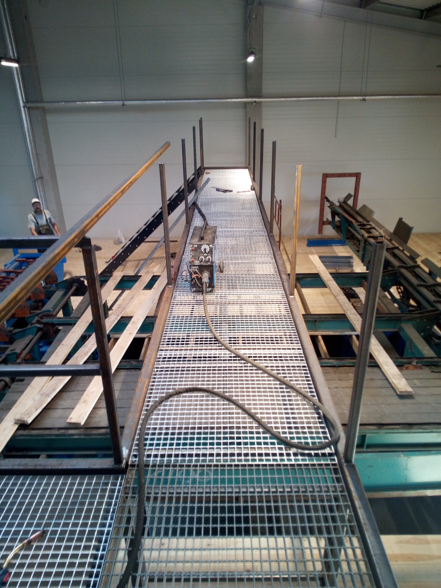 Konstrukcja stalowa pomostu roboczego w hali produkcyjnej, z widocznym spawaczem w oddali i maszyną spawalniczą na kratownicy, elementy konstrukcyjne w trakcie montażu.