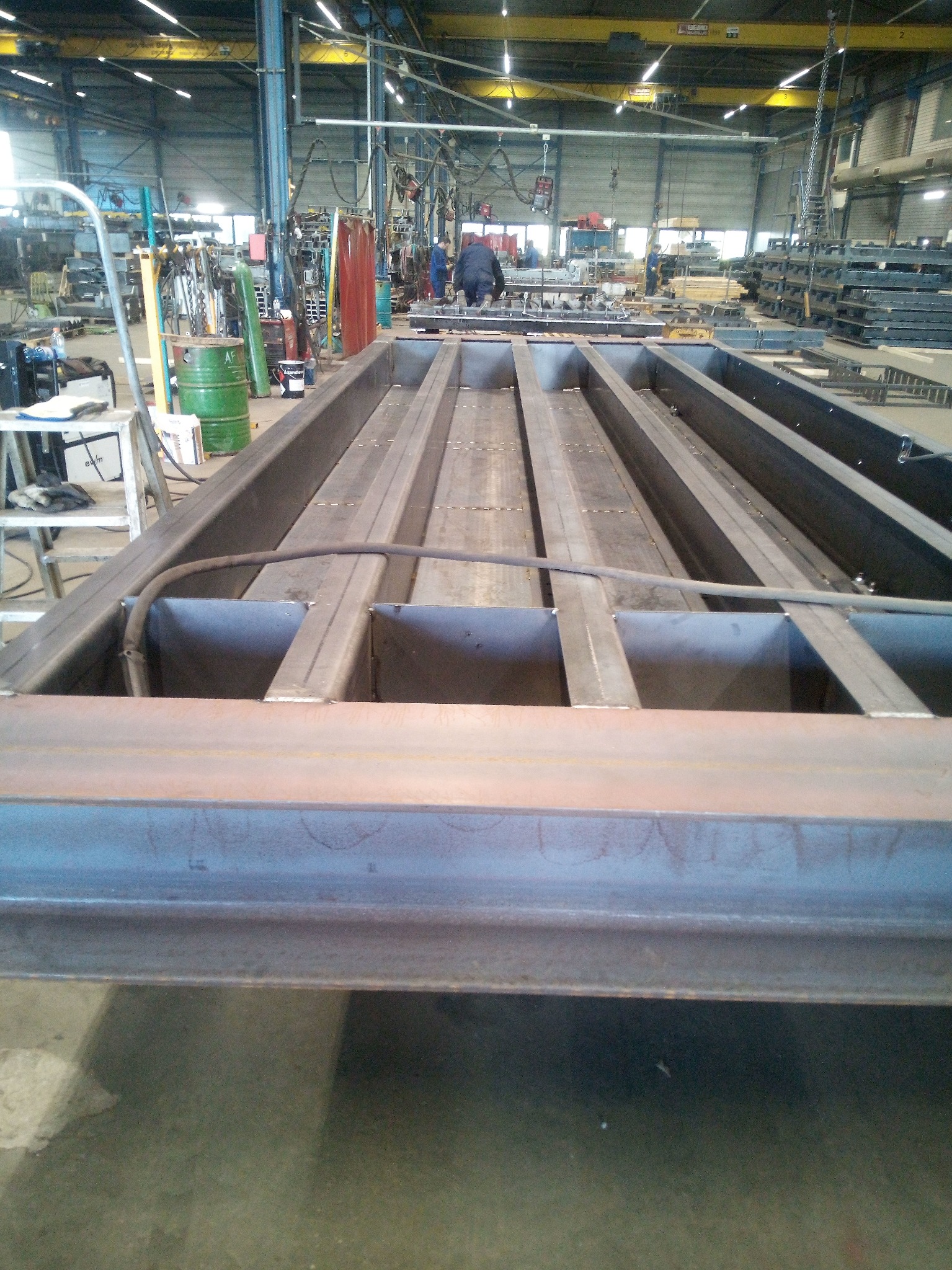 Konstrukcja stalowa platformy w hali produkcyjnej, widoczne spawy i elementy wzmacniające, w tle sprzęt spawalniczy i pracownik w kombinezonie.