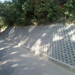 Ukończona, szara ściana oporowa z ażurowych elementów betonowych, zabezpieczająca skarpę. W tle zieleń i fragment ulicy z pracującym robotnikiem.