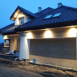 Nowoczesny dom murowany z drewnianą elewacją, oświetleniem zewnętrznym i ciemną dachówką, widziany o zmierzchu, teren wokół domu w trakcie prac wykończeniowych.