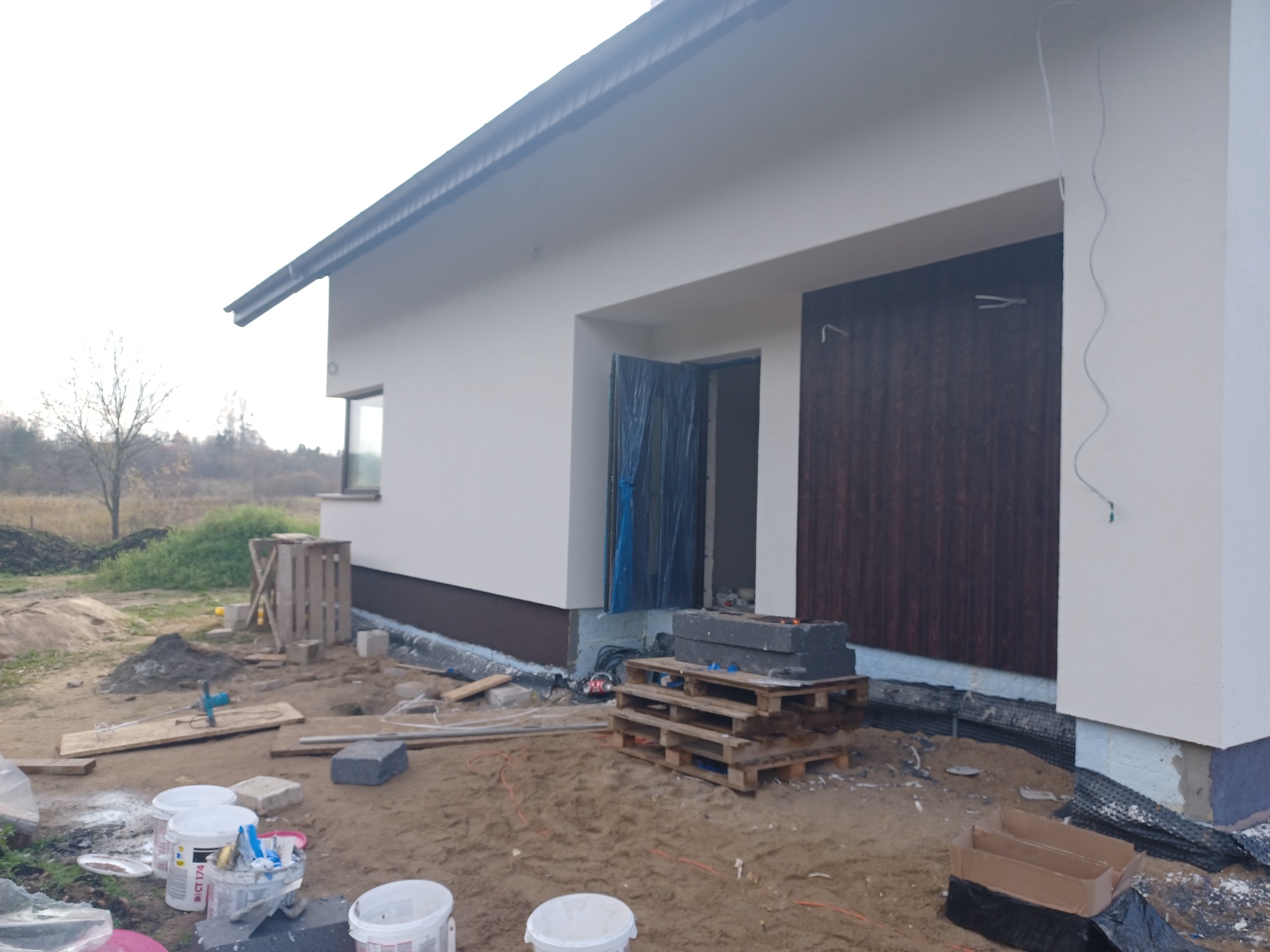 Niedokończony, parterowy budynek z brązowymi drzwiami garażowymi, widoczne materiały budowlane, palety i puste pojemniki po farbach na piaszczystym podłożu.
