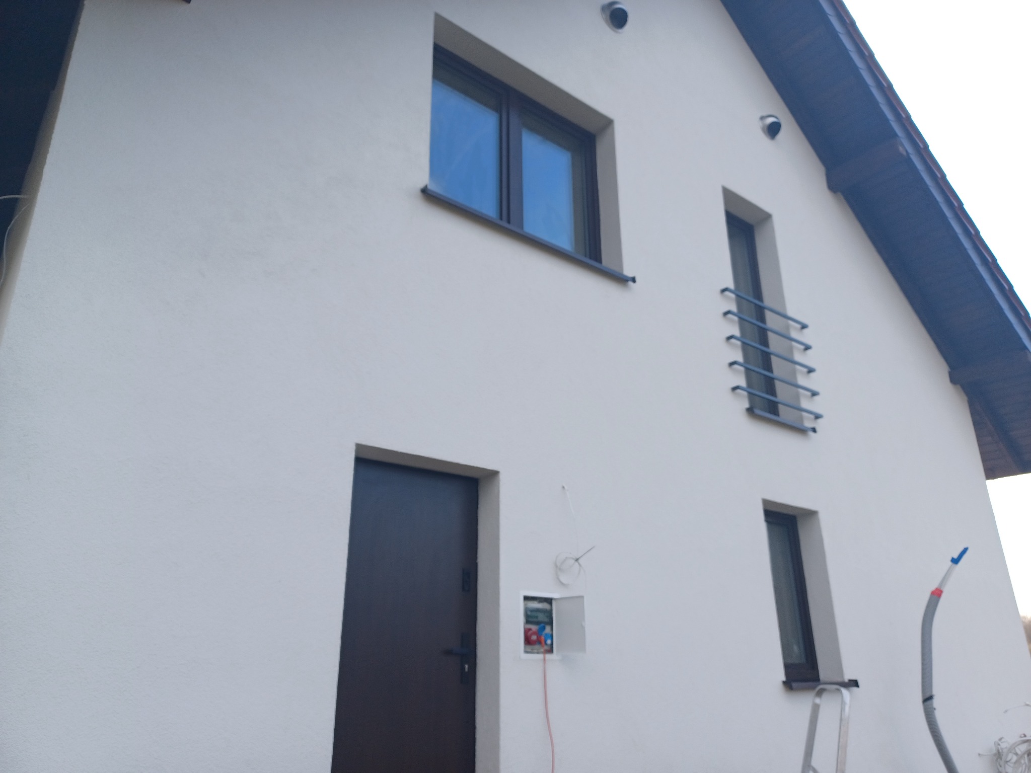 Świeżo otynkowany, dwukondygnacyjny dom z ciemnymi oknami, drzwiami i orynnowaniem, z widoczną skrzynką elektryczną, przewodem i drabiną opartą o ścianę.